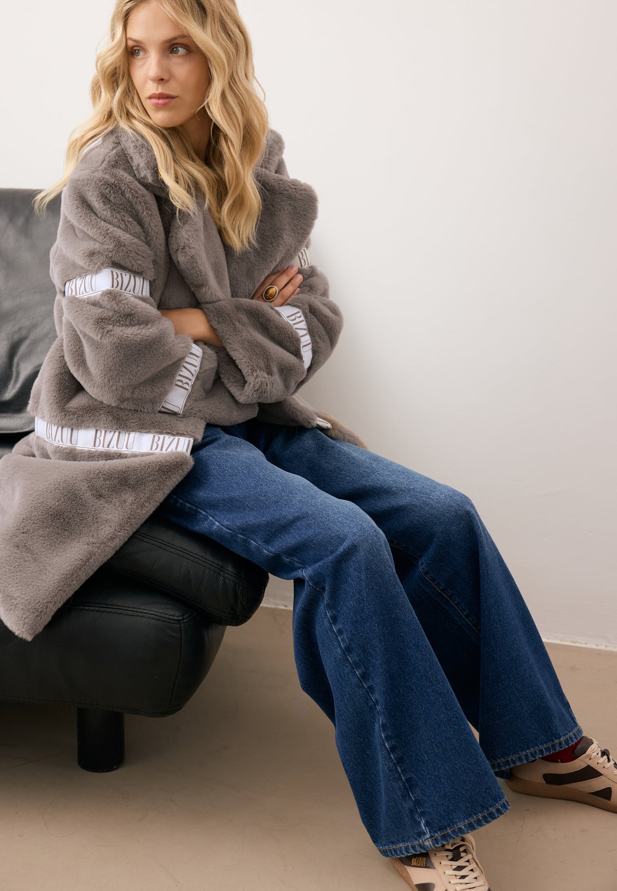 Model sitting on a sofa with arms crossed over her chest, wearing the taupe FLORENCE fur coat and navy LEYA jeans.