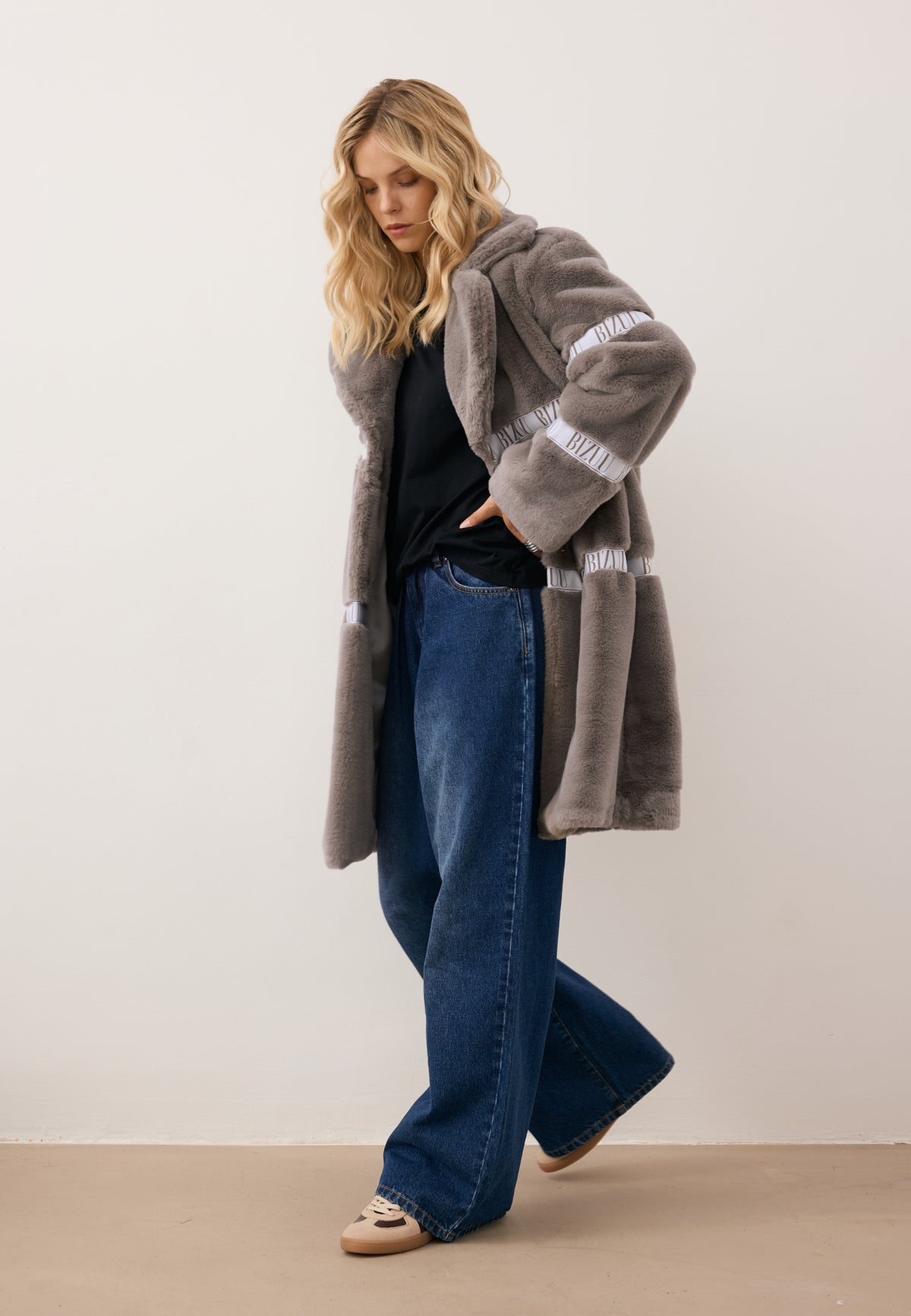 Model stepping forward with one hand on her hip, dressed in the taupe FLORENCE faux fur coat with branded tape.
