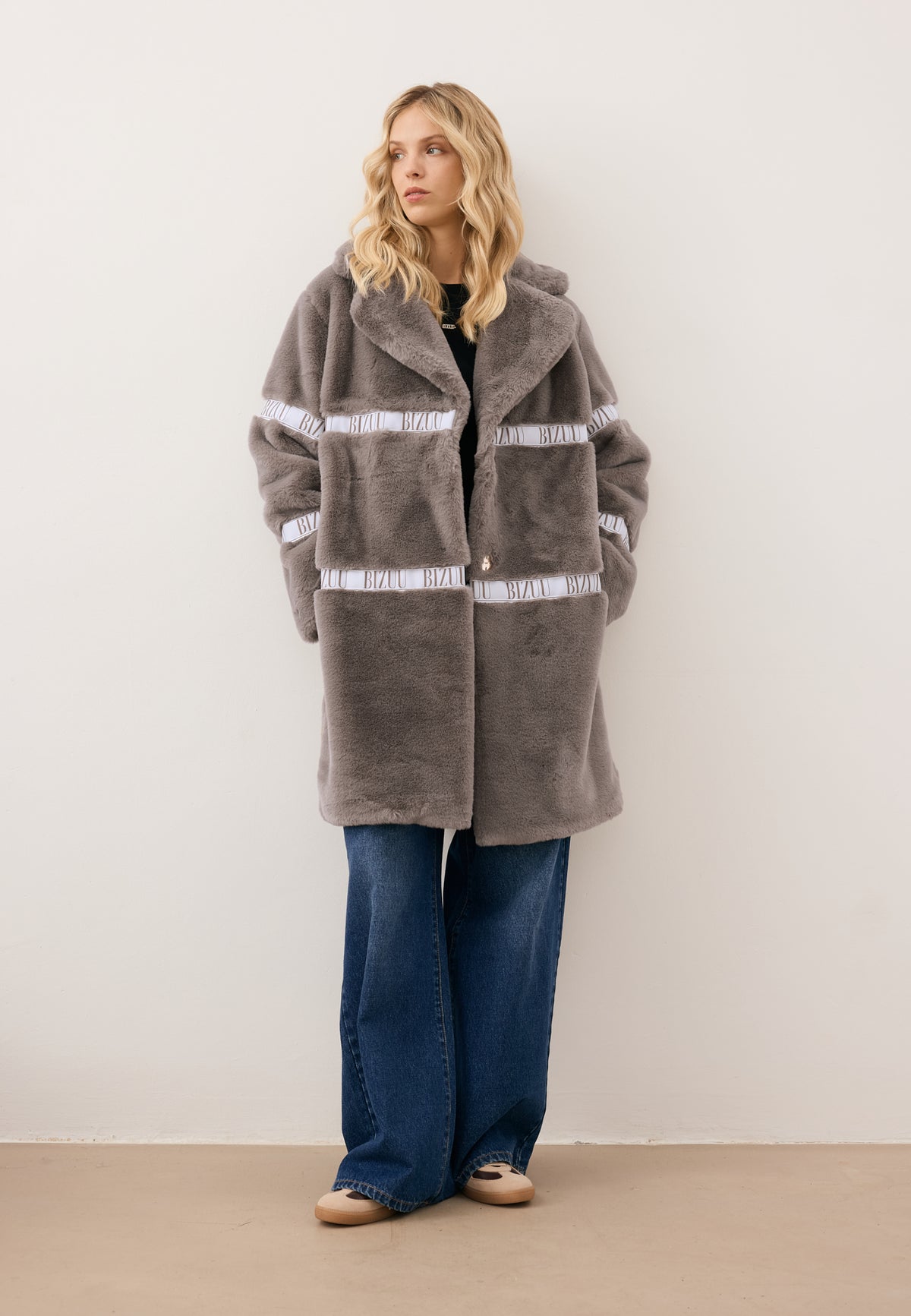 Model looking to the side with hands in the pockets of the taupe FLORENCE fur coat with logo detail.
