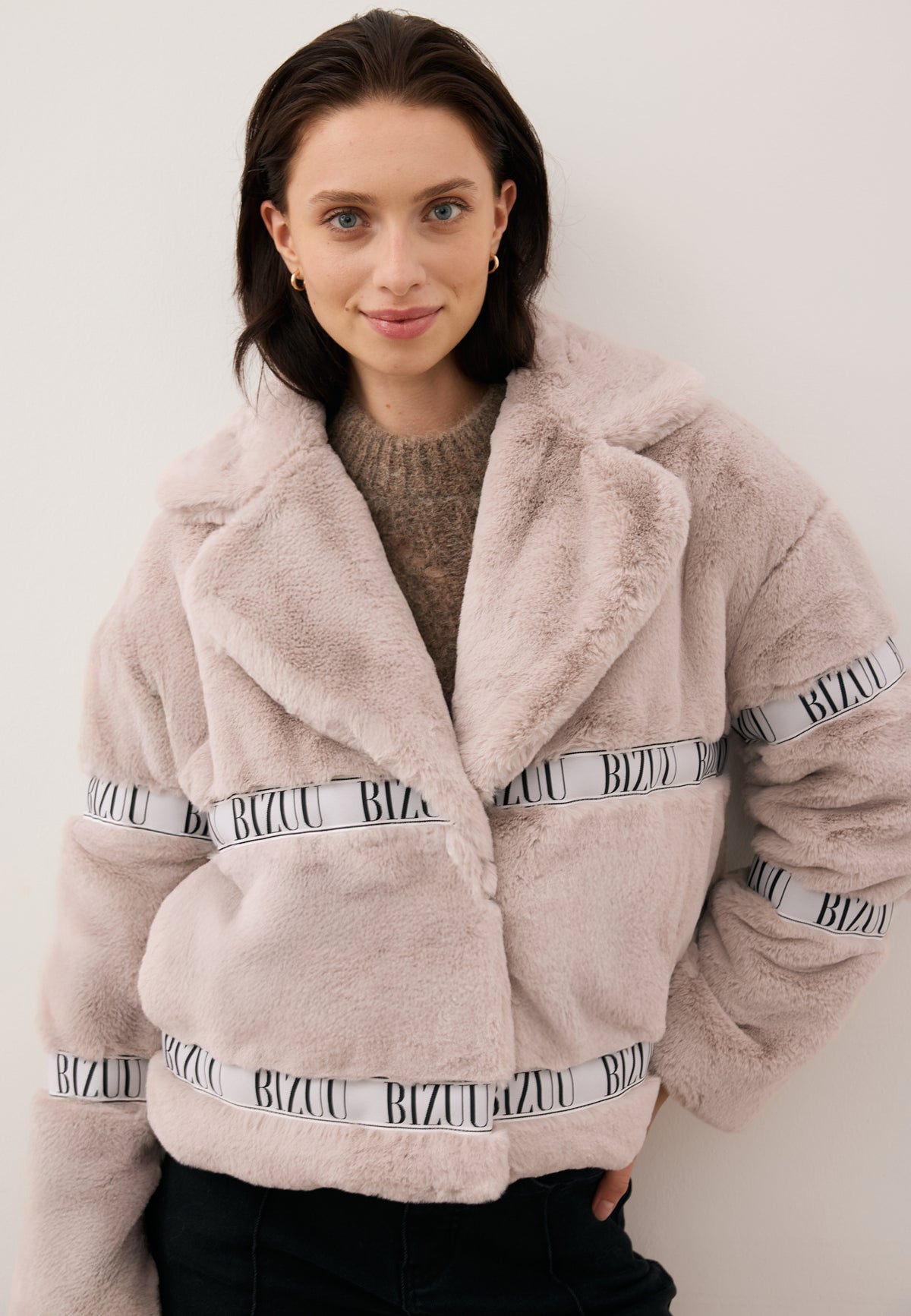 Model with one hand behind her back, wearing the beige short GREGORIA faux fur coat with logo tape.