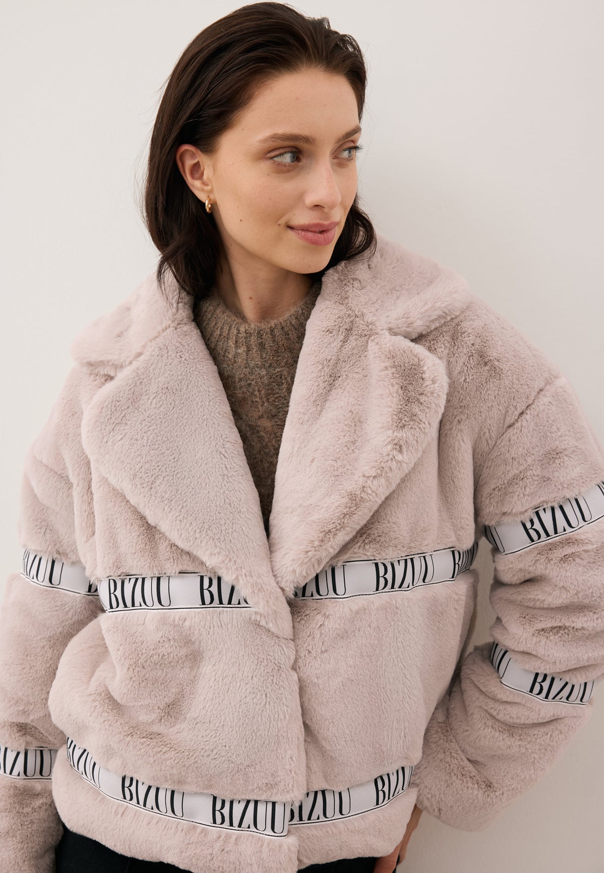 Model looking to the side, dressed in the short beige GREGORIA faux fur coat with branded stripe.
