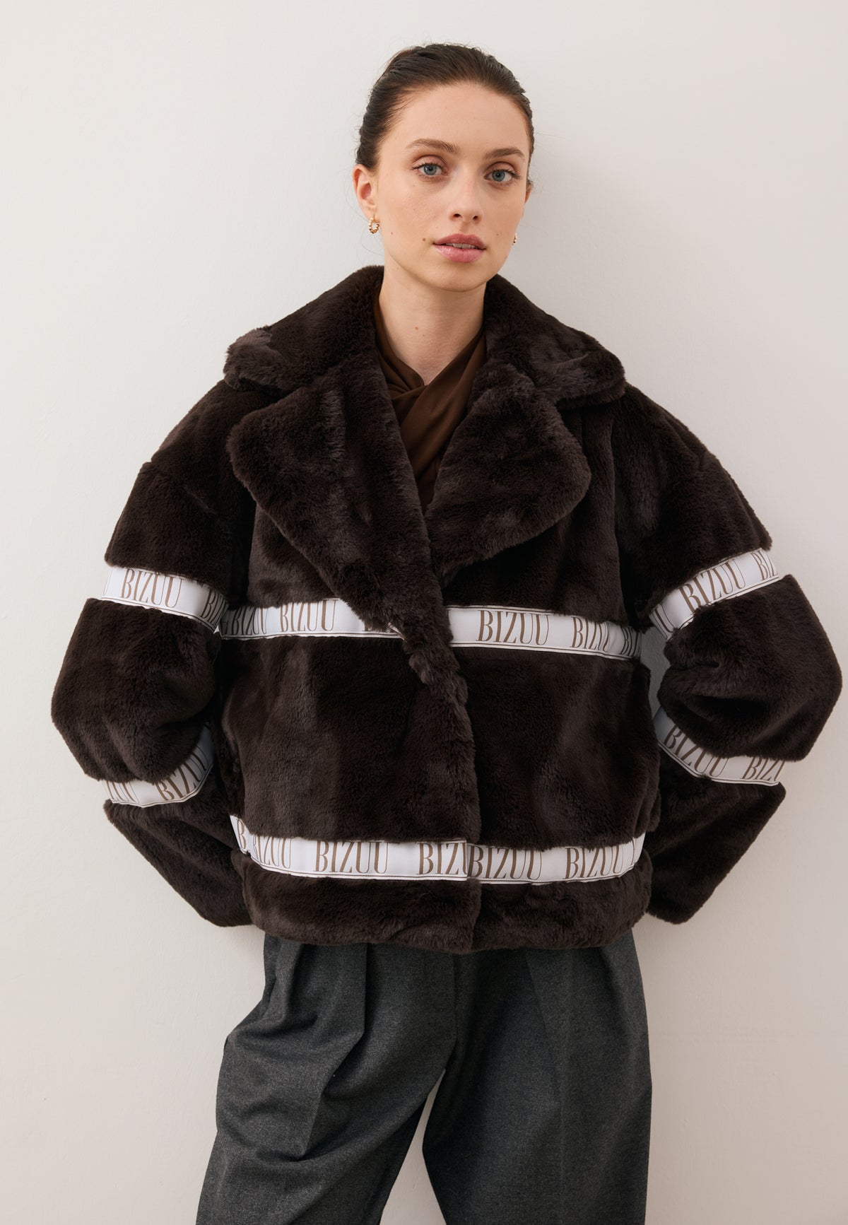 Model with hands behind her back, wearing the boxy short brown GREGORIA faux fur coat with logo tape.
