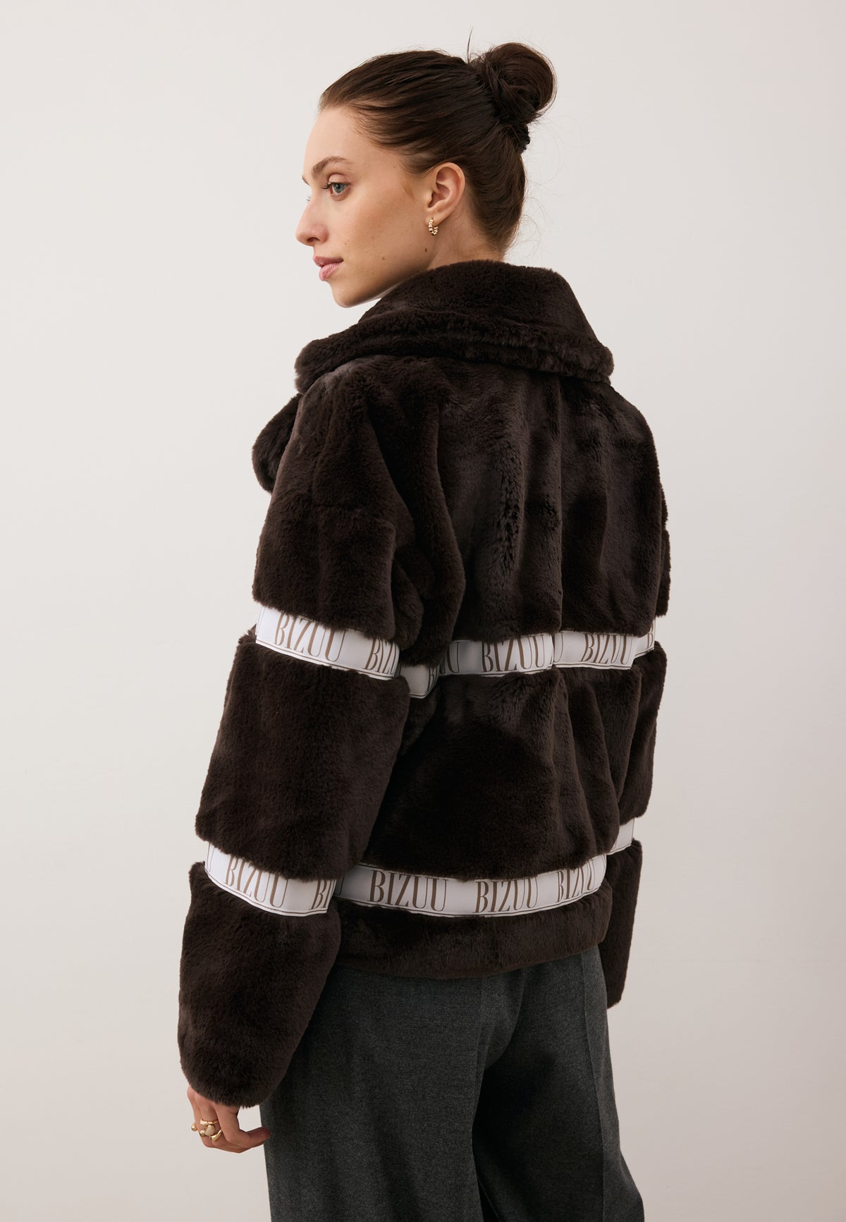 Model in a side view, dressed in the short GREGORIA faux fur coat with branded stripe.
