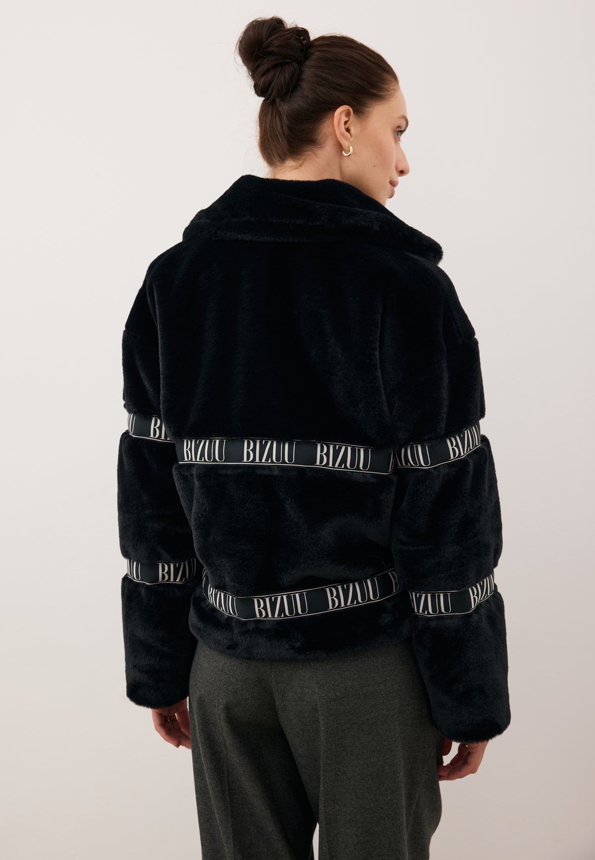 Model standing with her back to the camera in the short black GREGORIA faux fur coat with logo tape.
