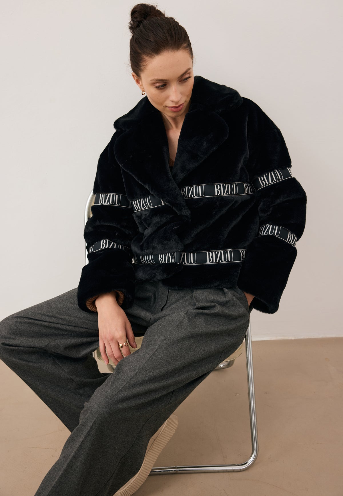 Smiling model with hands in BANOS trouser pockets, wearing the black short GREGORIA faux fur coat with logo tape.