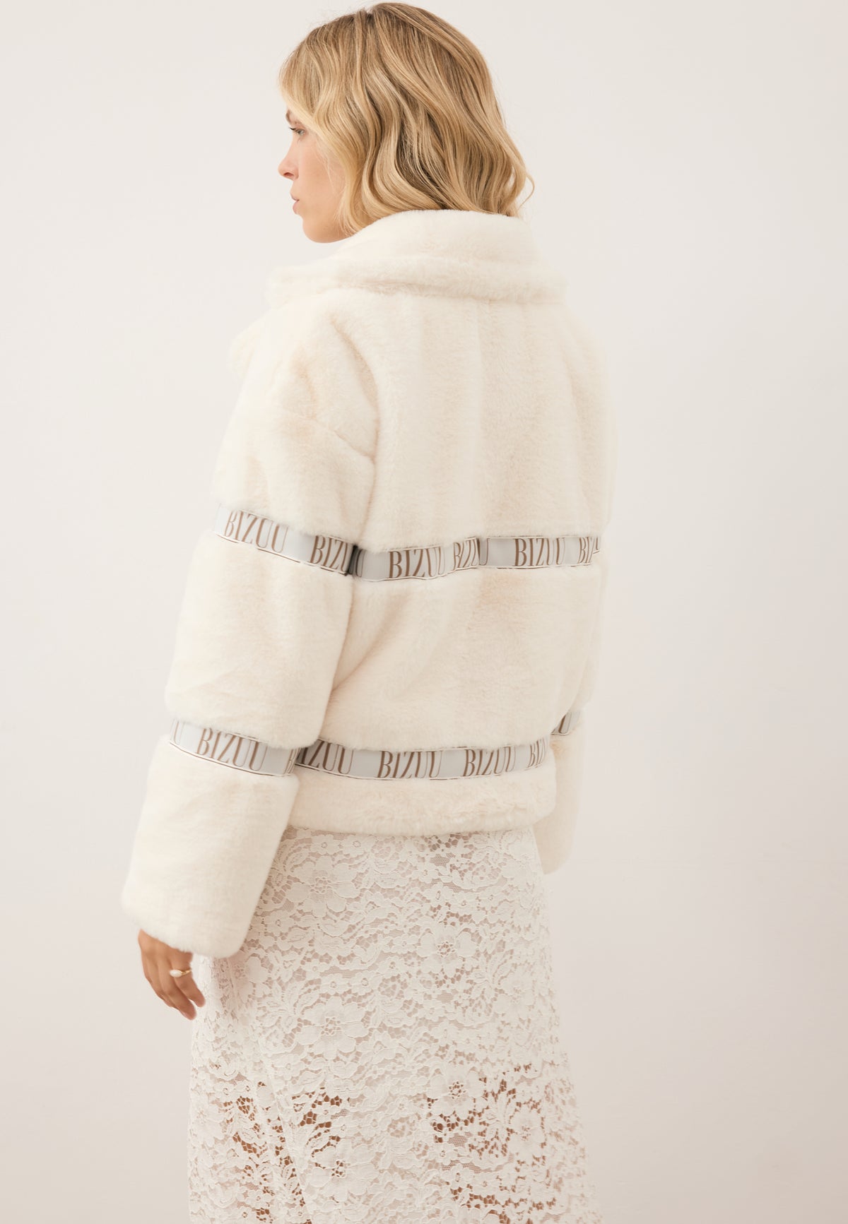 Model standing with her back to the camera, wearing the short cream GREGORIA faux fur coat with logo tape.