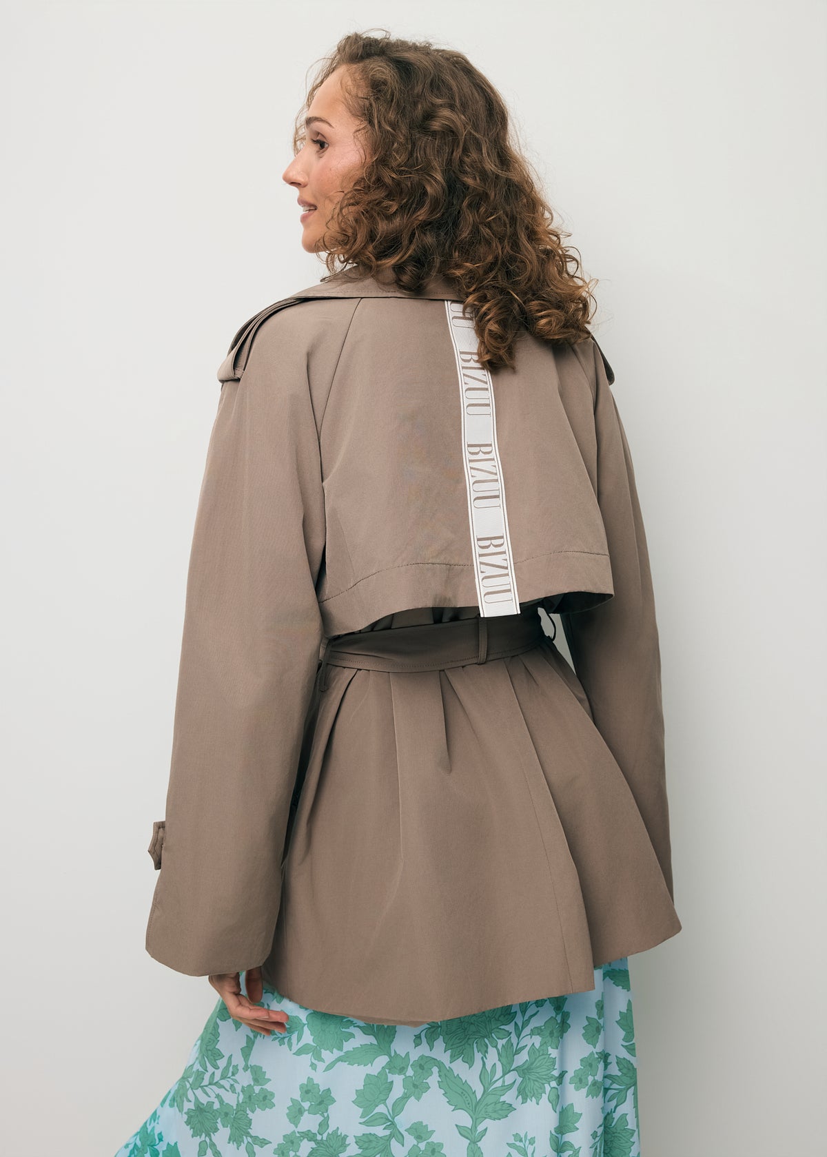 A model standing with her back to the camera, wearing a short RACHY trench coat with a logo tape detail on the back.