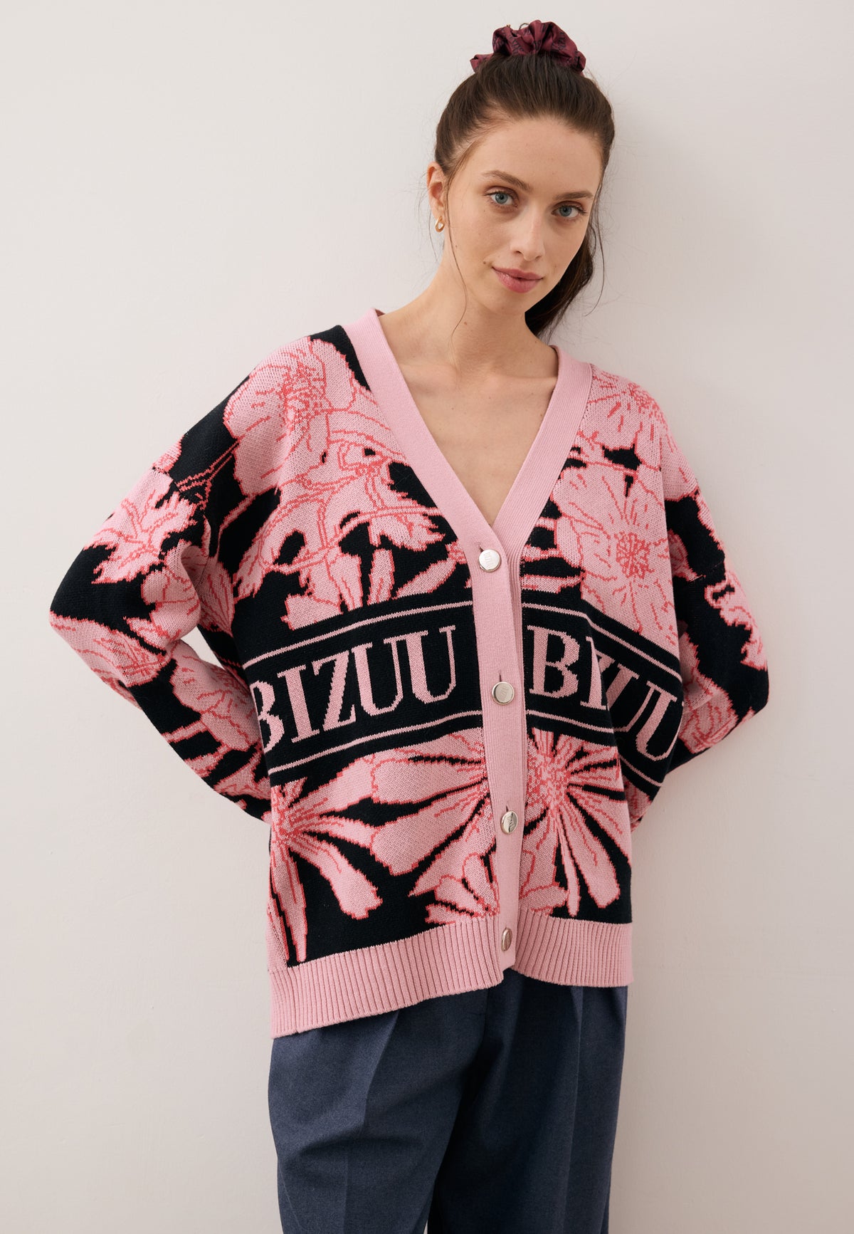 Model with her hands behind her back, looking straight ahead, dressed in the pink and black HYLES cardigan with branded buttons.