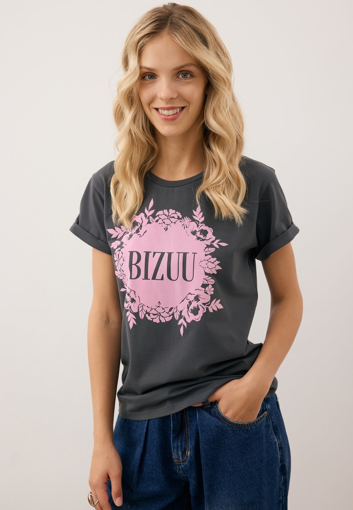 Smiling model with one hand in the pocket of LEYA trousers, wearing the graphite IOLA T-shirt with pink appliqué.