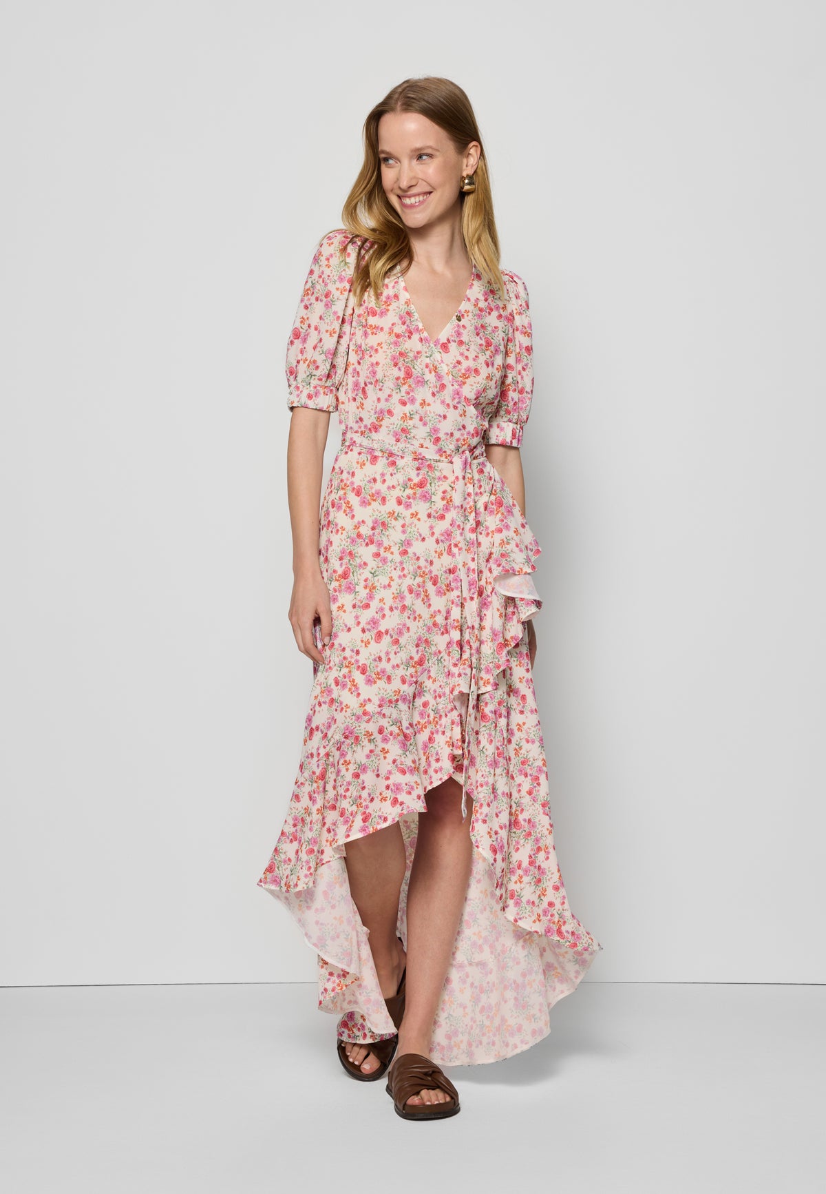 Model in asymmetrical envelope dress in pink flowers with ruffle and waist belt ISLAY, wearing brown flip-flops.