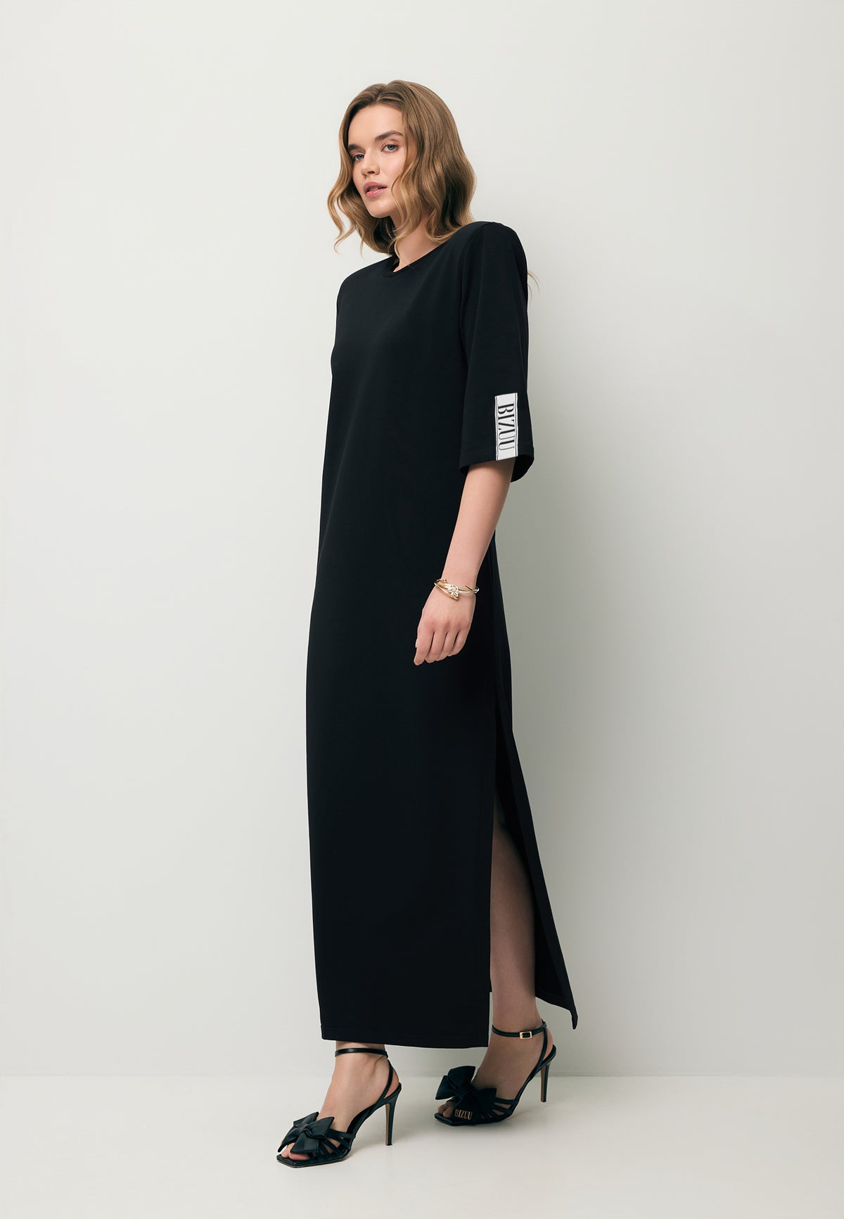 A model stepping forward wearing the JULISSA knitted maxi dress with a logo tape on the sleeve.