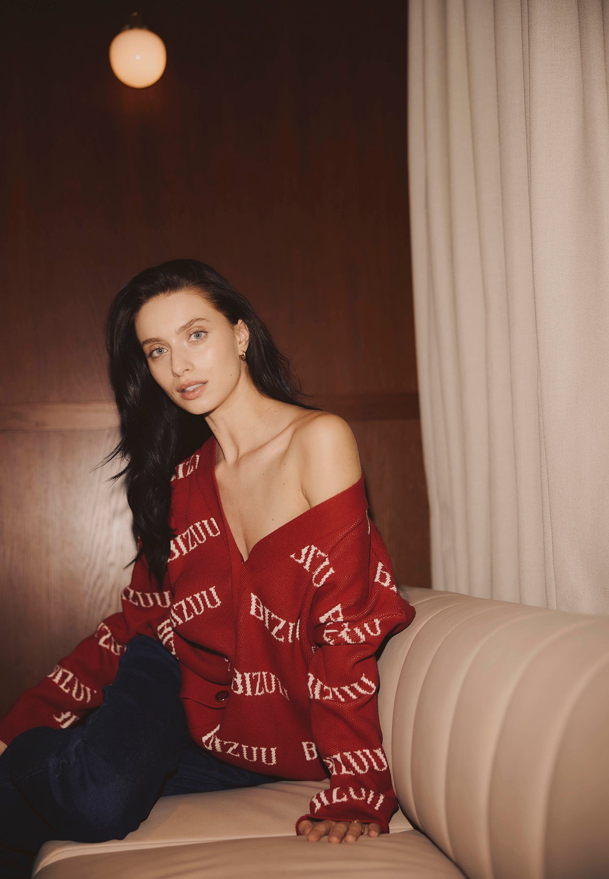Model sitting on a sofa wearing the burgundy KYLE cardigan with a cream logo.