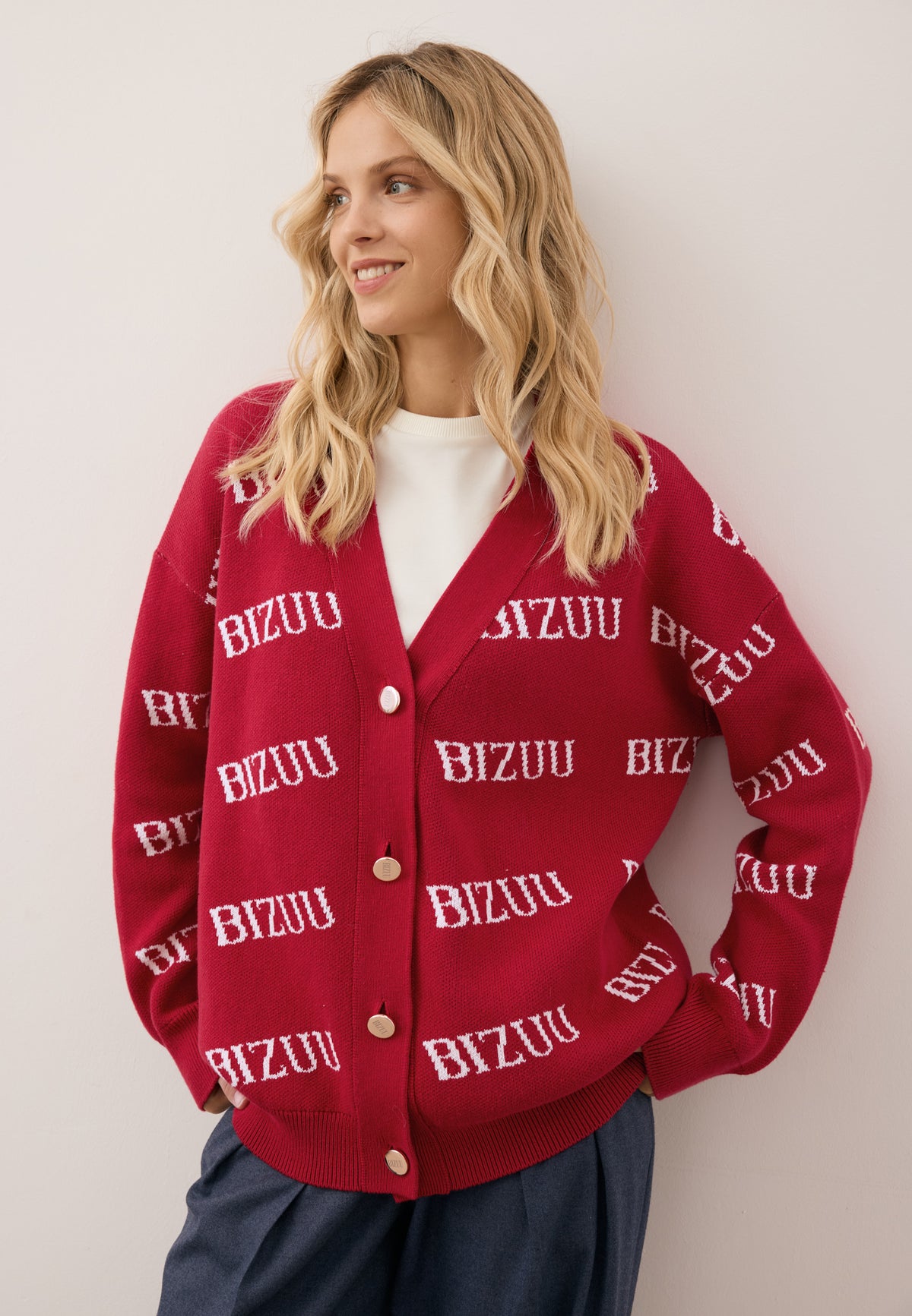 Close-up of the model leaning against a wall wearing the burgundy KYLE cardigan with a logo pattern, fastened with golden buttons.