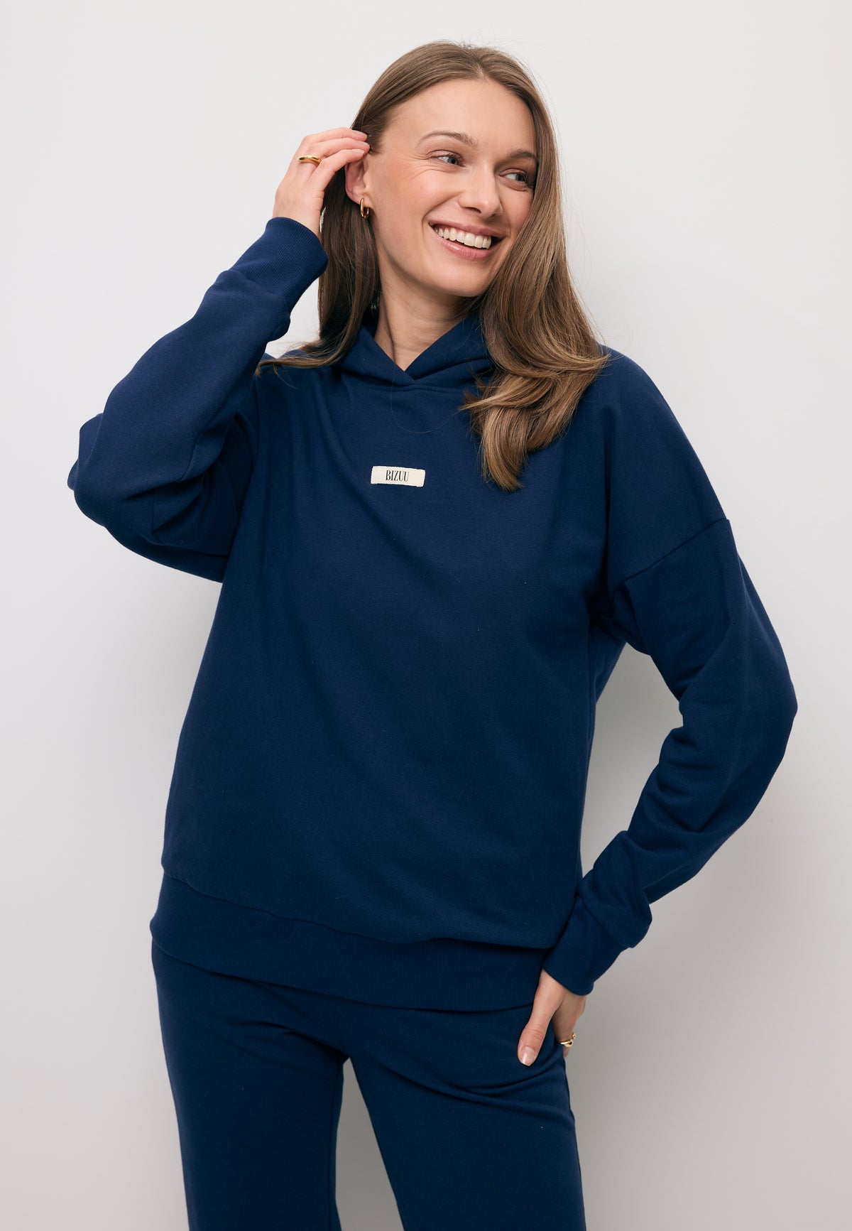 Close-up of a smiling model adjusting her hair, wearing a navy LINDI hoodie with a branded label on the front.