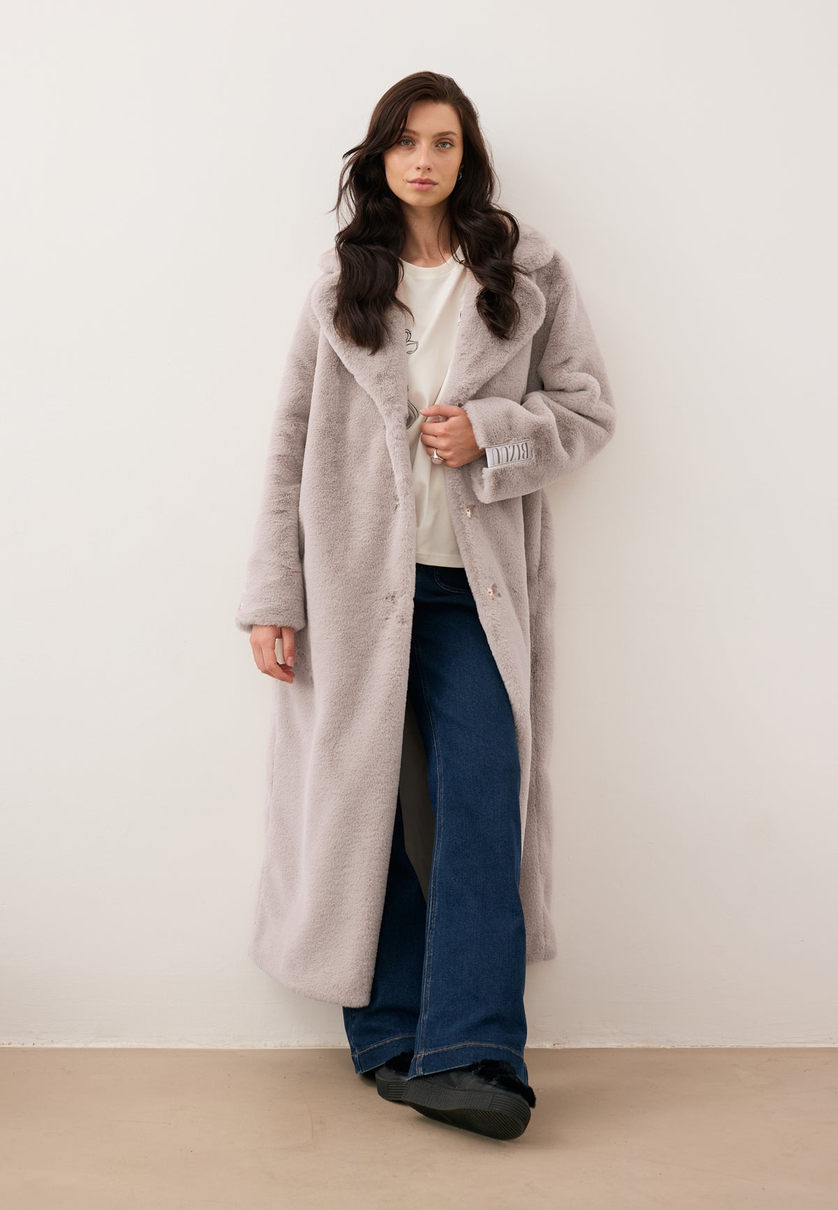 Model leaning against a wall, holding the collar of a beige midi-length LIVORNO fur coat with a logo on the sleeve.