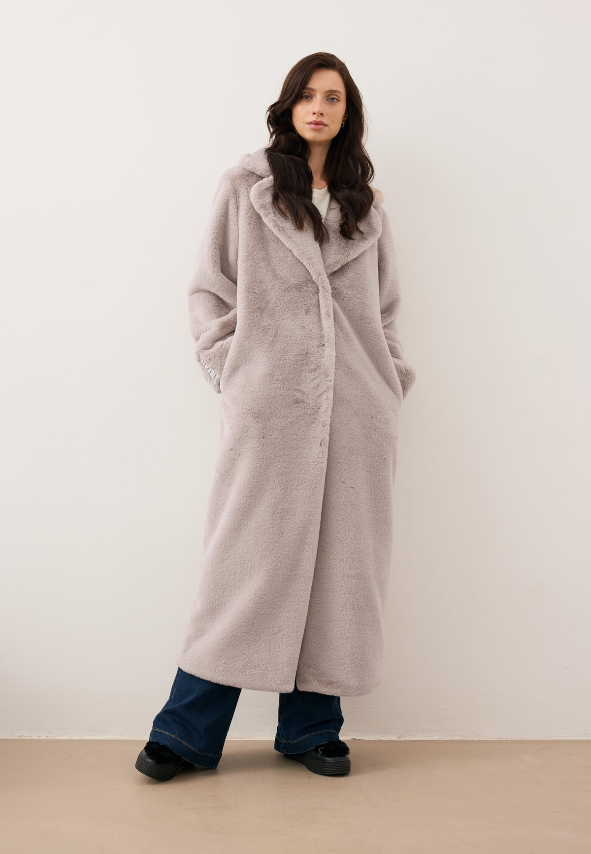 Model against a white wall in a beige, long LIVORNO fur coat with branded tape on the back and a logo on the sleeve.