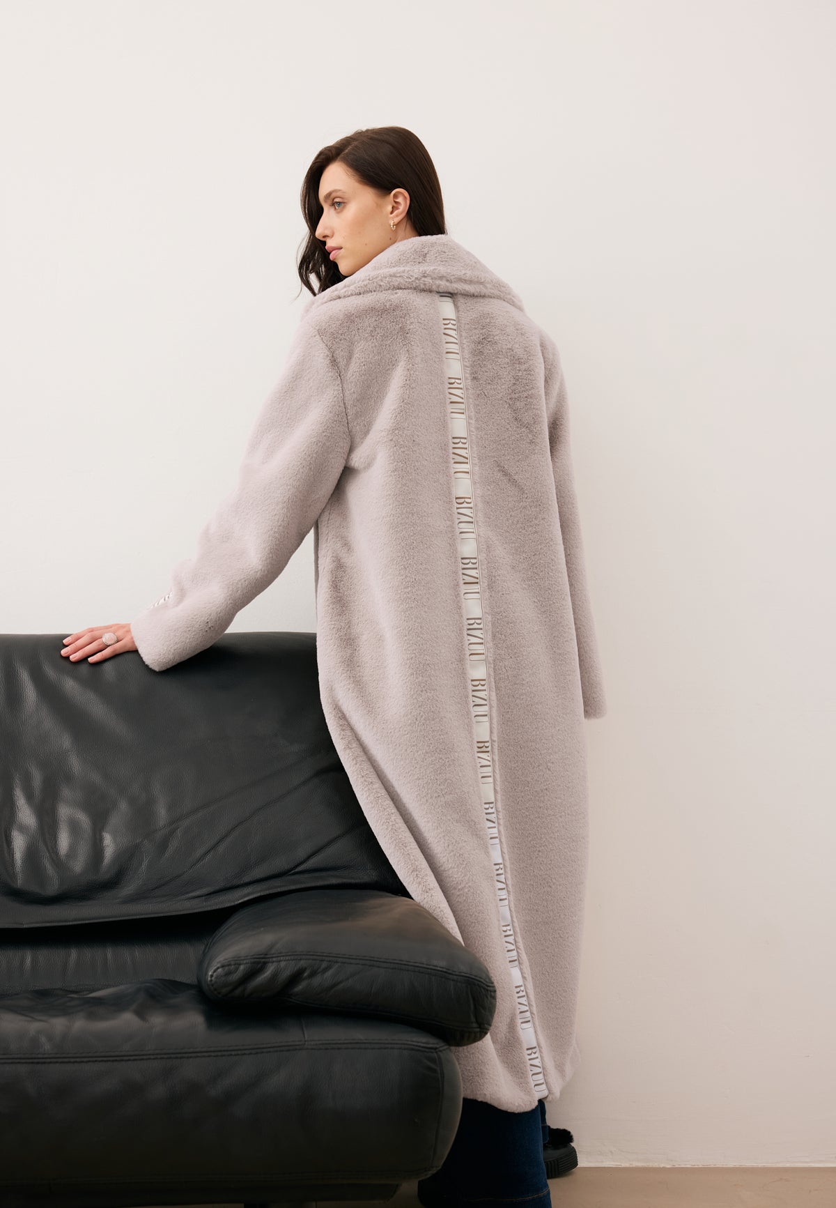 Model standing with her back turned, leaning against a sofa, wearing a long beige LIVORNO fur coat with a stripe on the back.