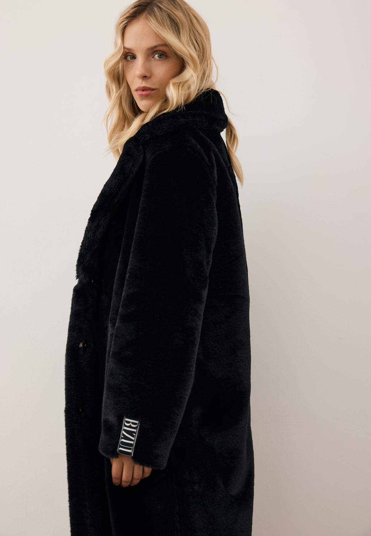 Model standing sideways wearing a black, long LIVORNO fur coat with a logo on the sleeve and branded tape on the back.