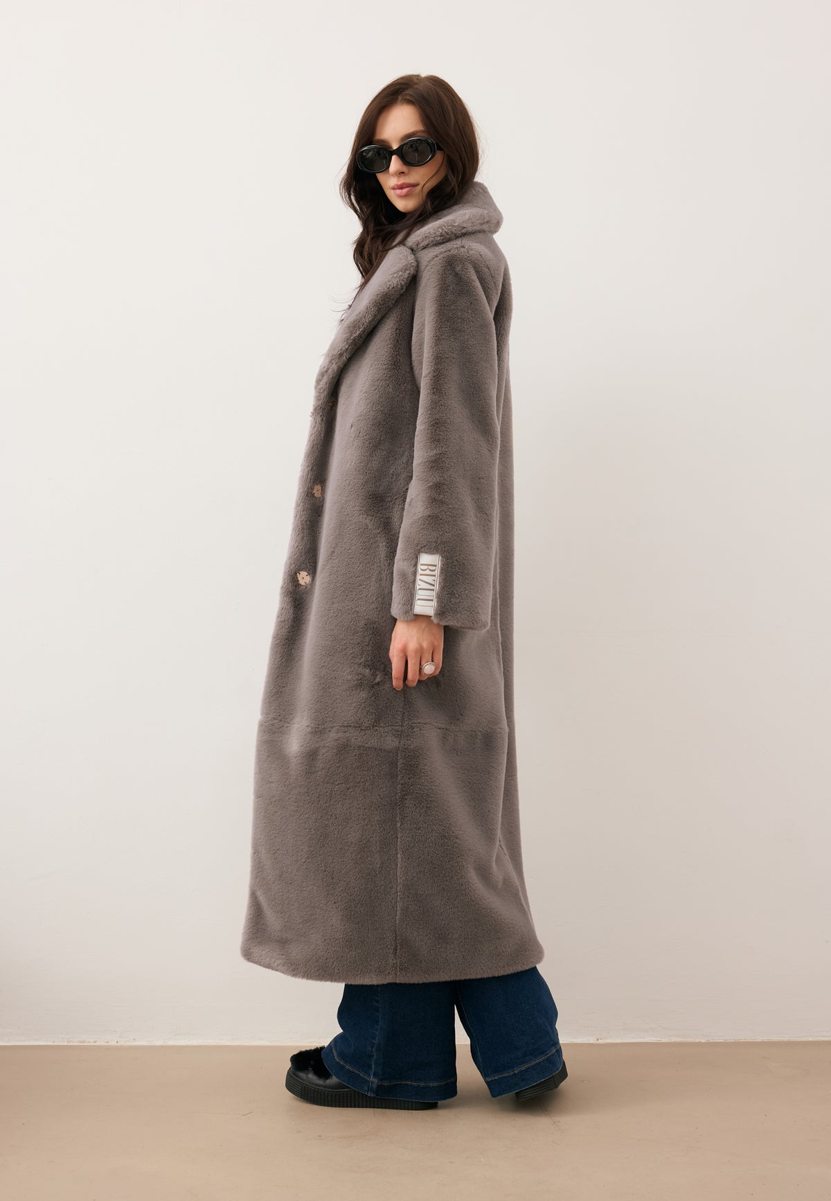Model in side view, wearing the long taupe LIVORNO faux fur coat.