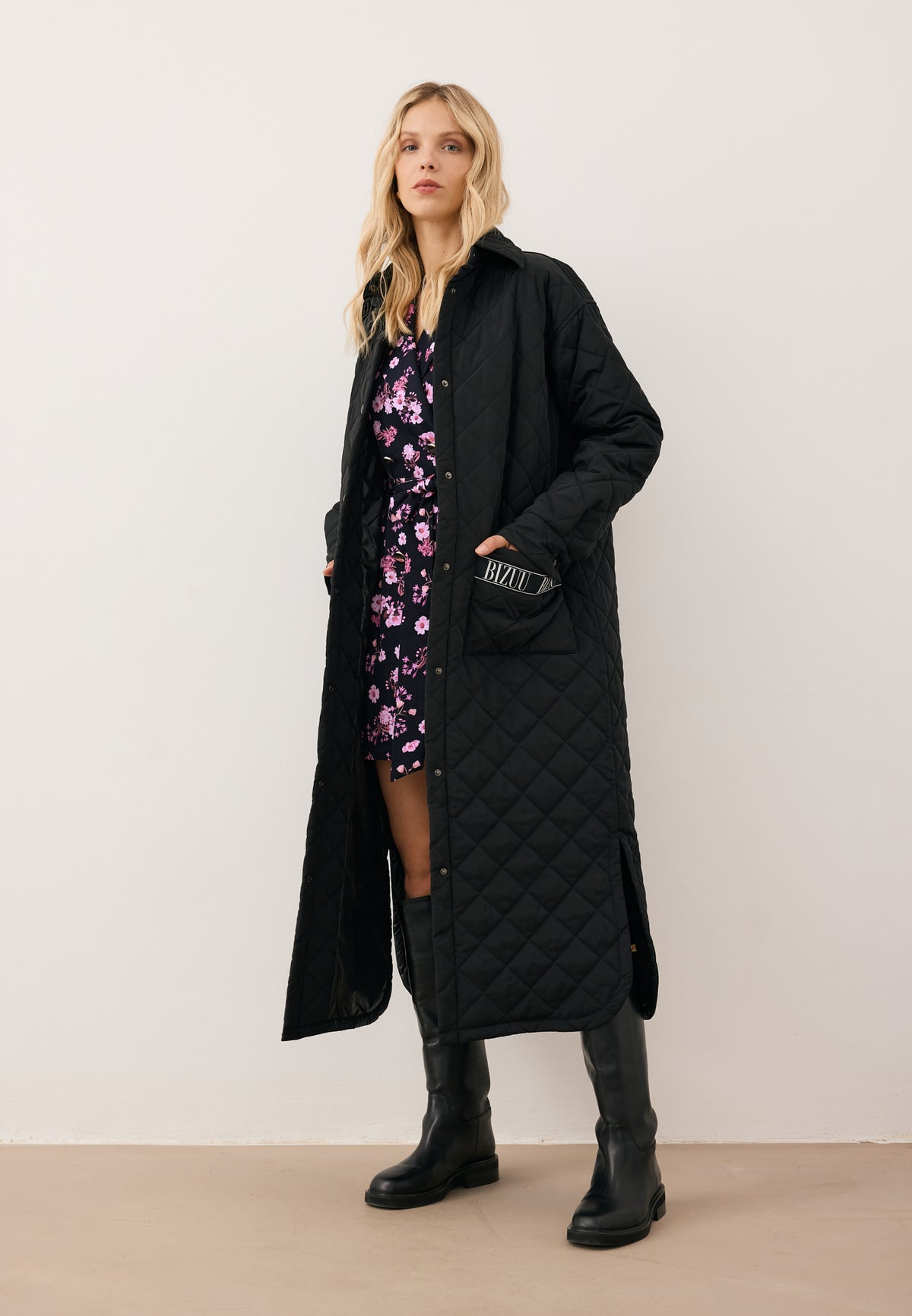 Model with one hand in the pocket of the MAREVIA quilted coat with logo detail on the pocket, styled with the GREGORIA floral waist-tie dress underneath.
