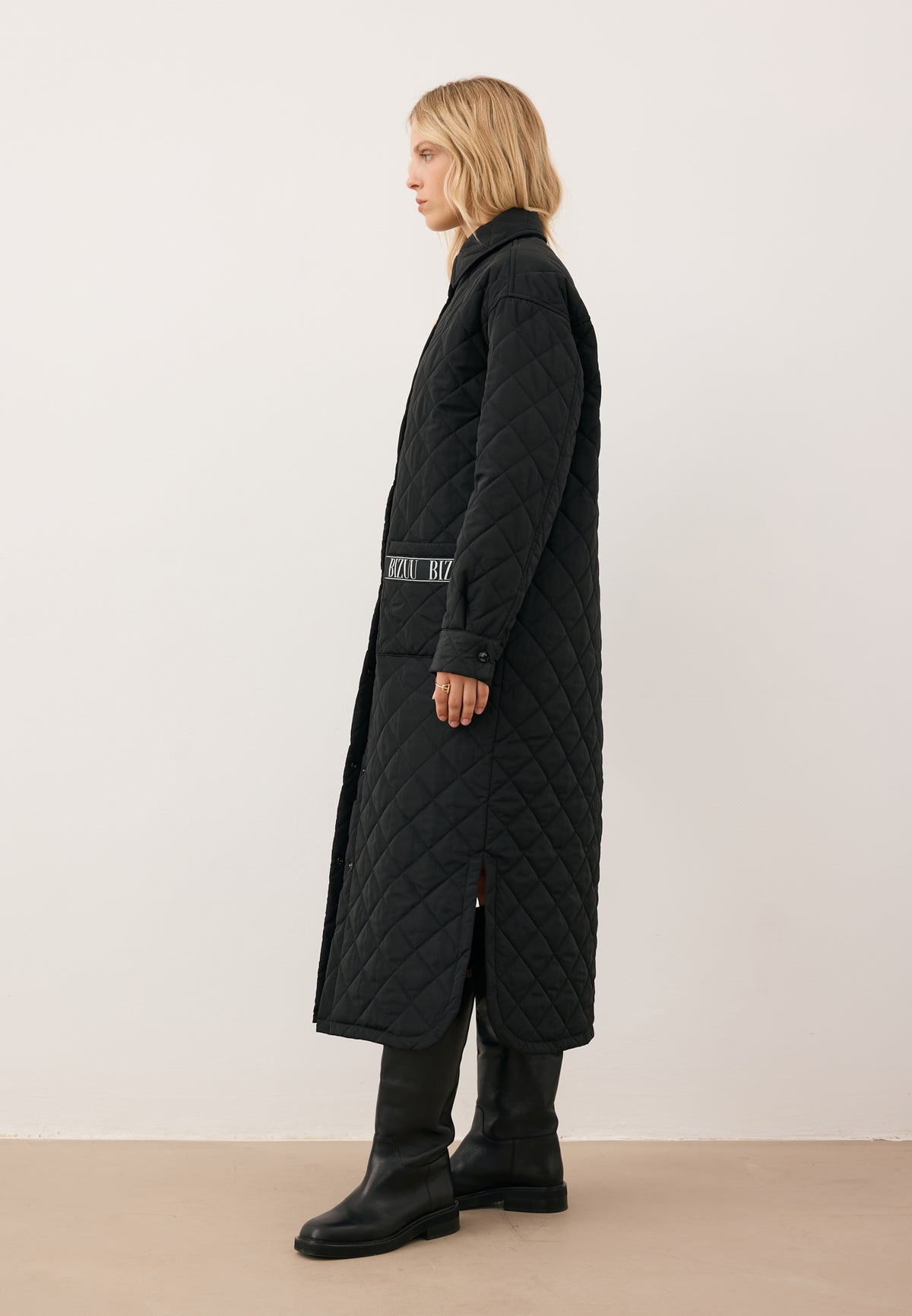 Model standing in profile, wearing the MAREVIA quilted coat with branded tape detail on the pocket.