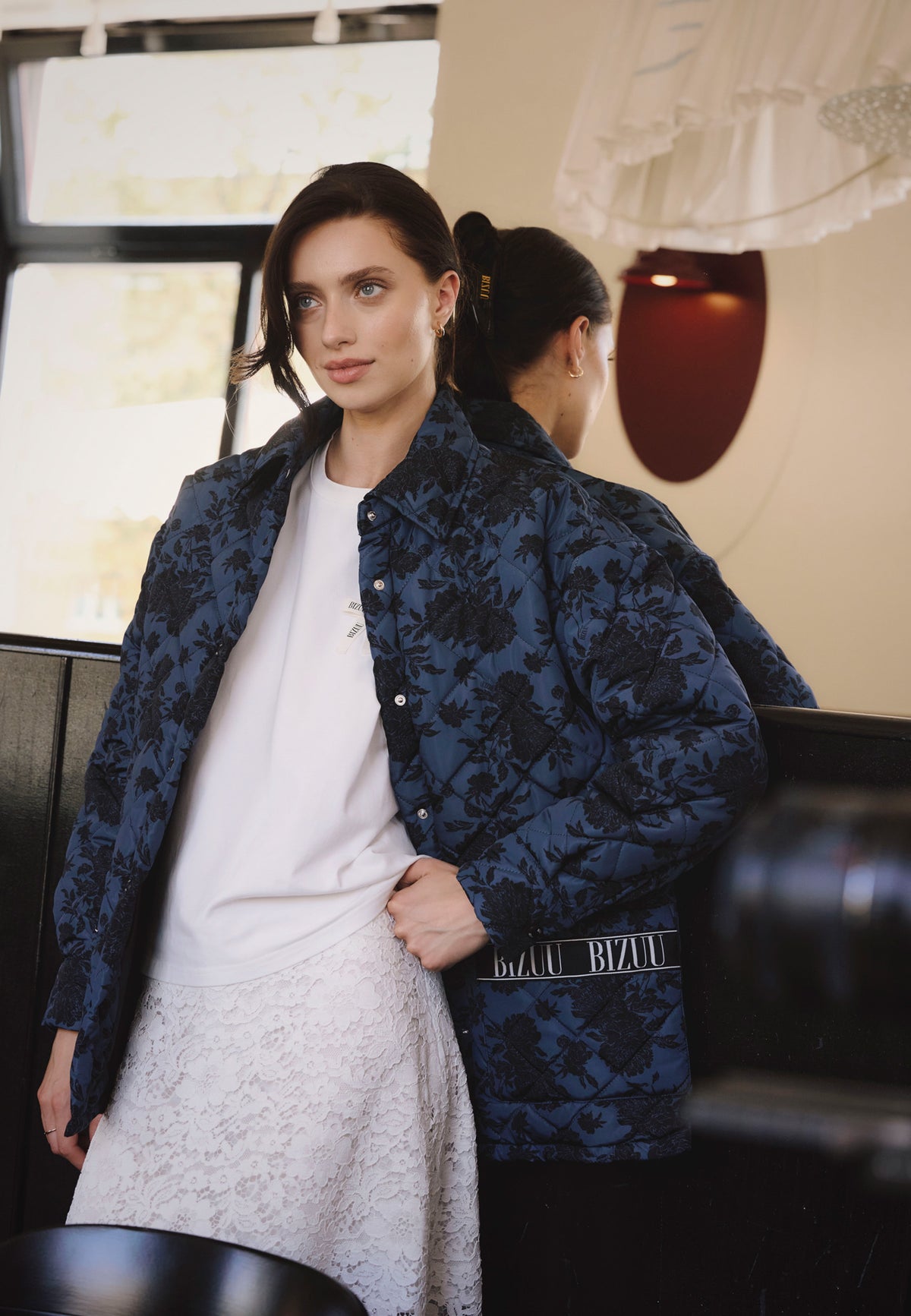 A model leaning against a wall wearing a white TISHALLE T-shirt, a white MILKY lace skirt, and a MARIN printed jacket.