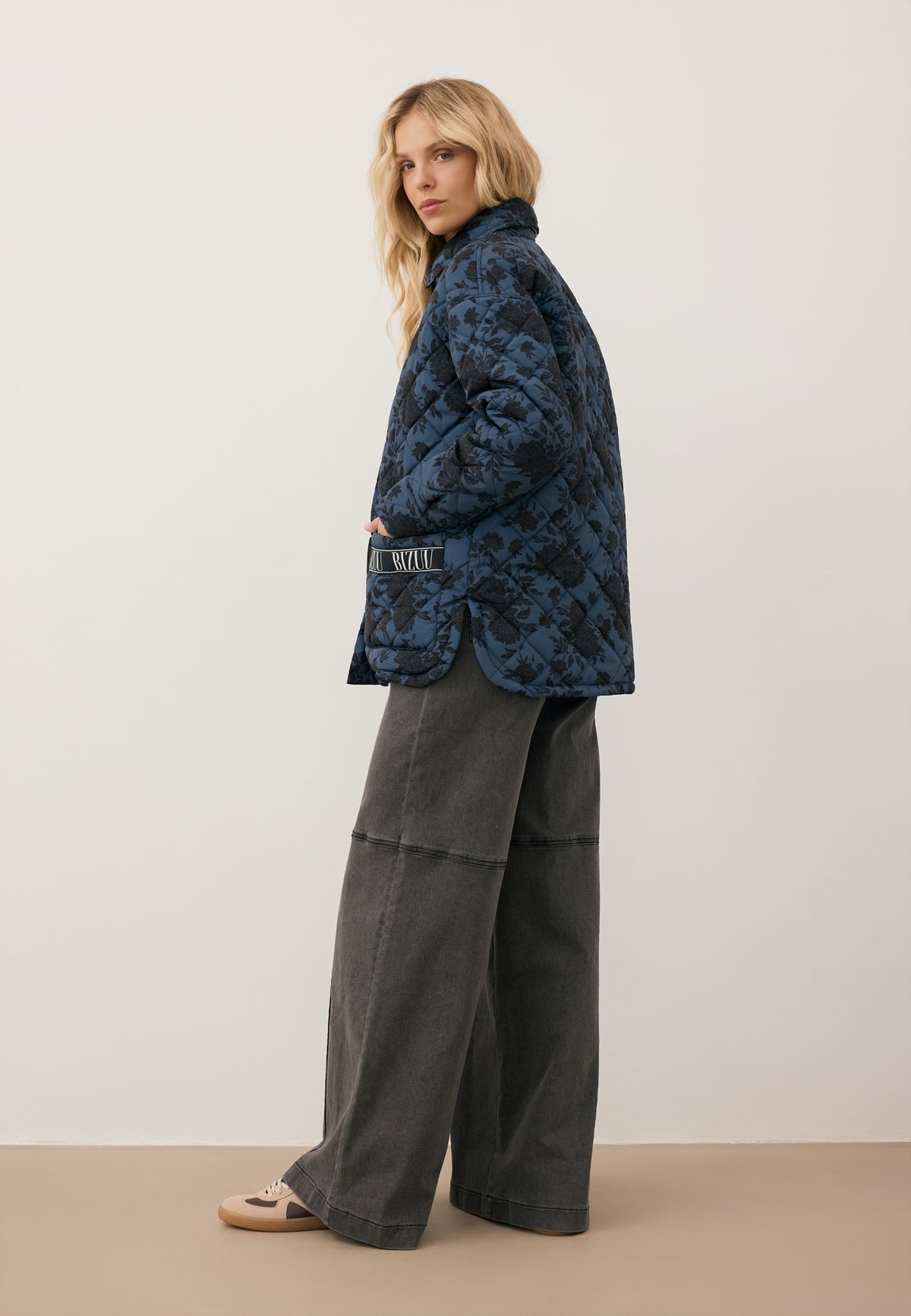 A model standing sideways against a white wall, wearing a MENIDNA jumpsuit and a MARIN quilted jacket.