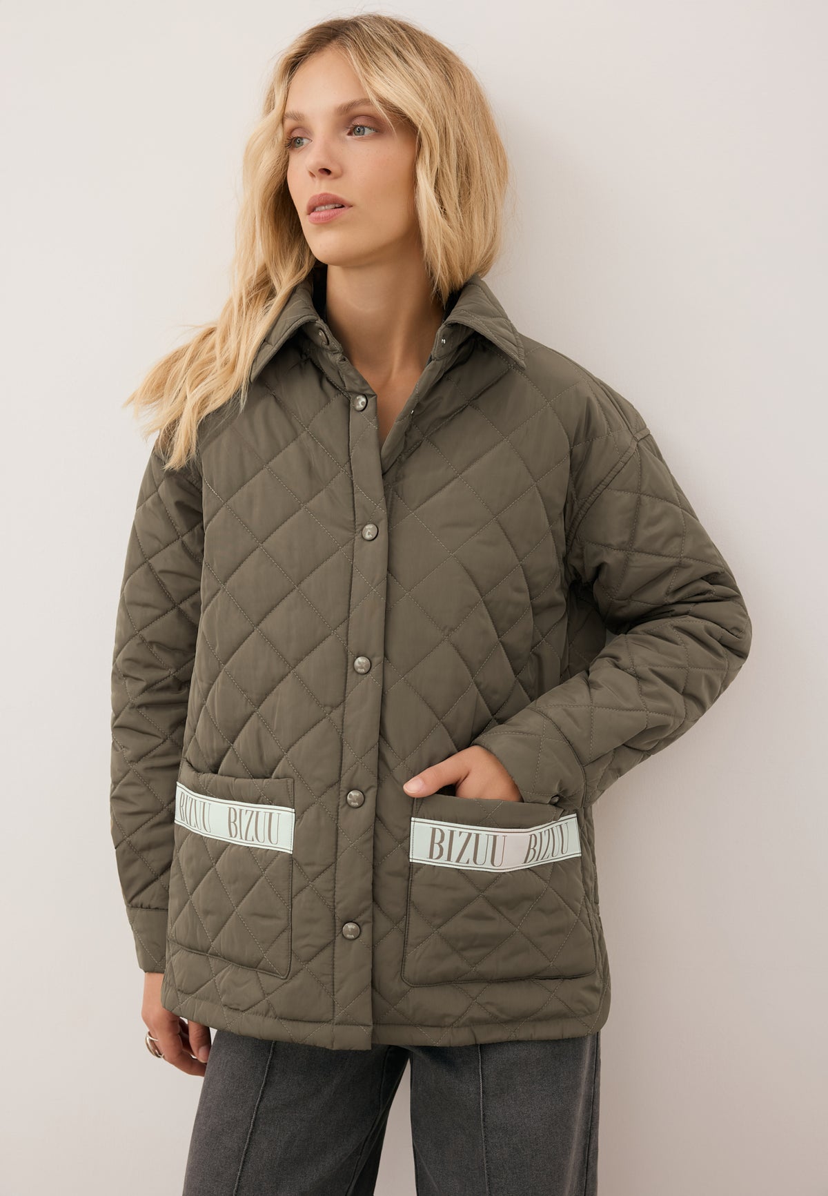 The model leaning against the wall has one hand in the pocket of her khaki, quilted MARIN jacket with a logo on the pockets.