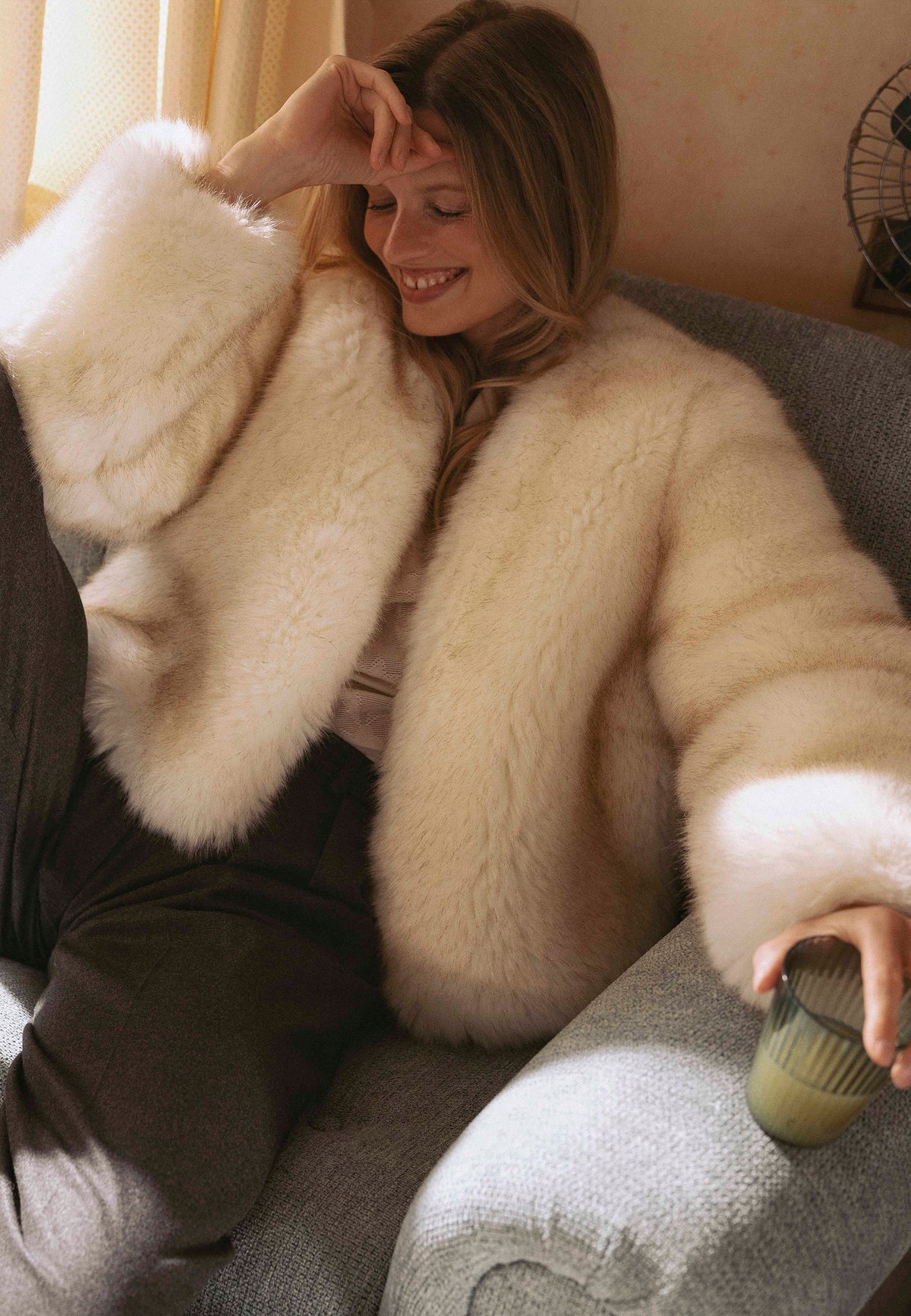 A smiling model sitting on a sofa wearing the short cream NOVARA faux fur coat.