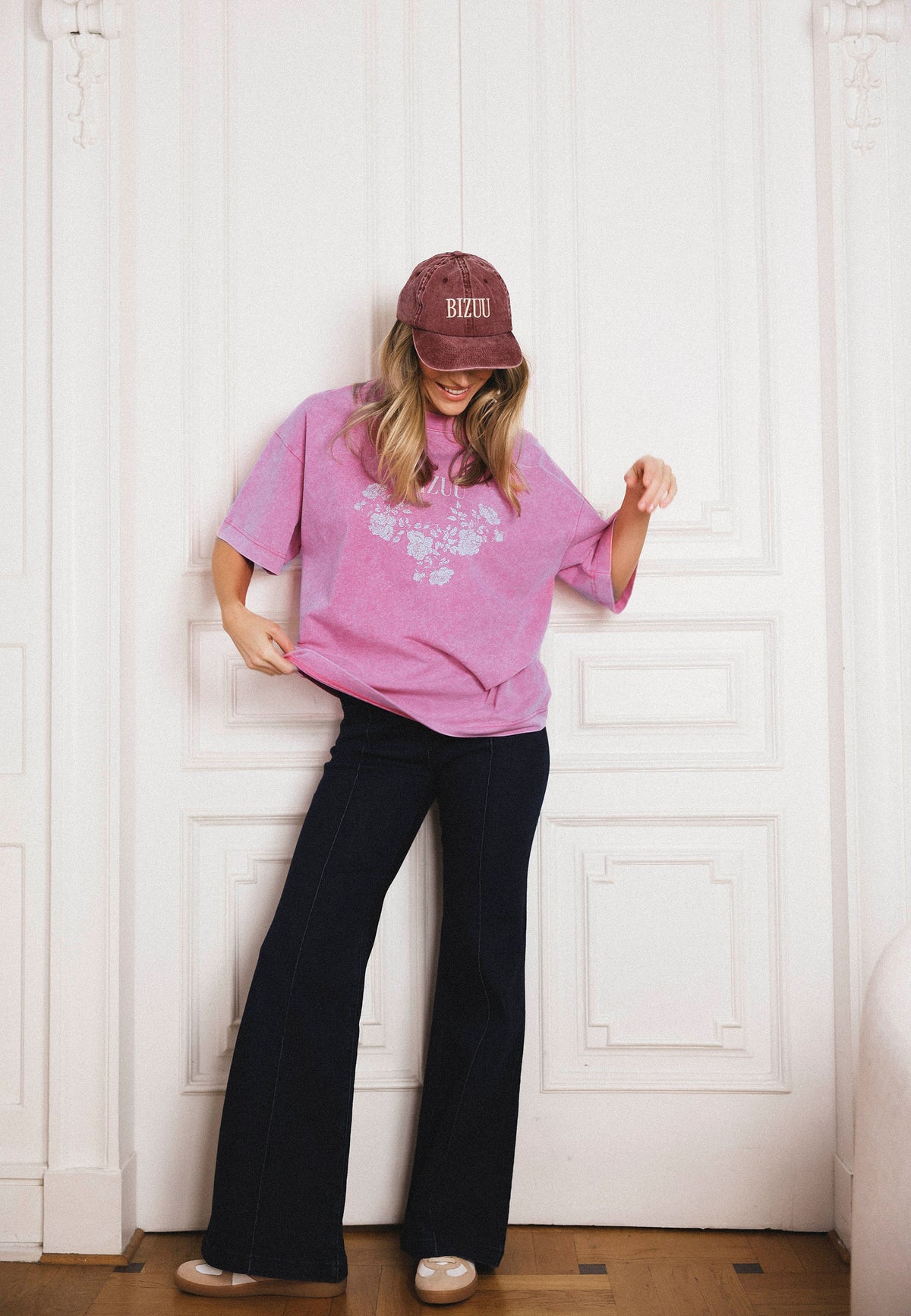 The model against a white door background, wearing a pink oversized PAPLYS T-shirt and BELINNA flared jeans.
