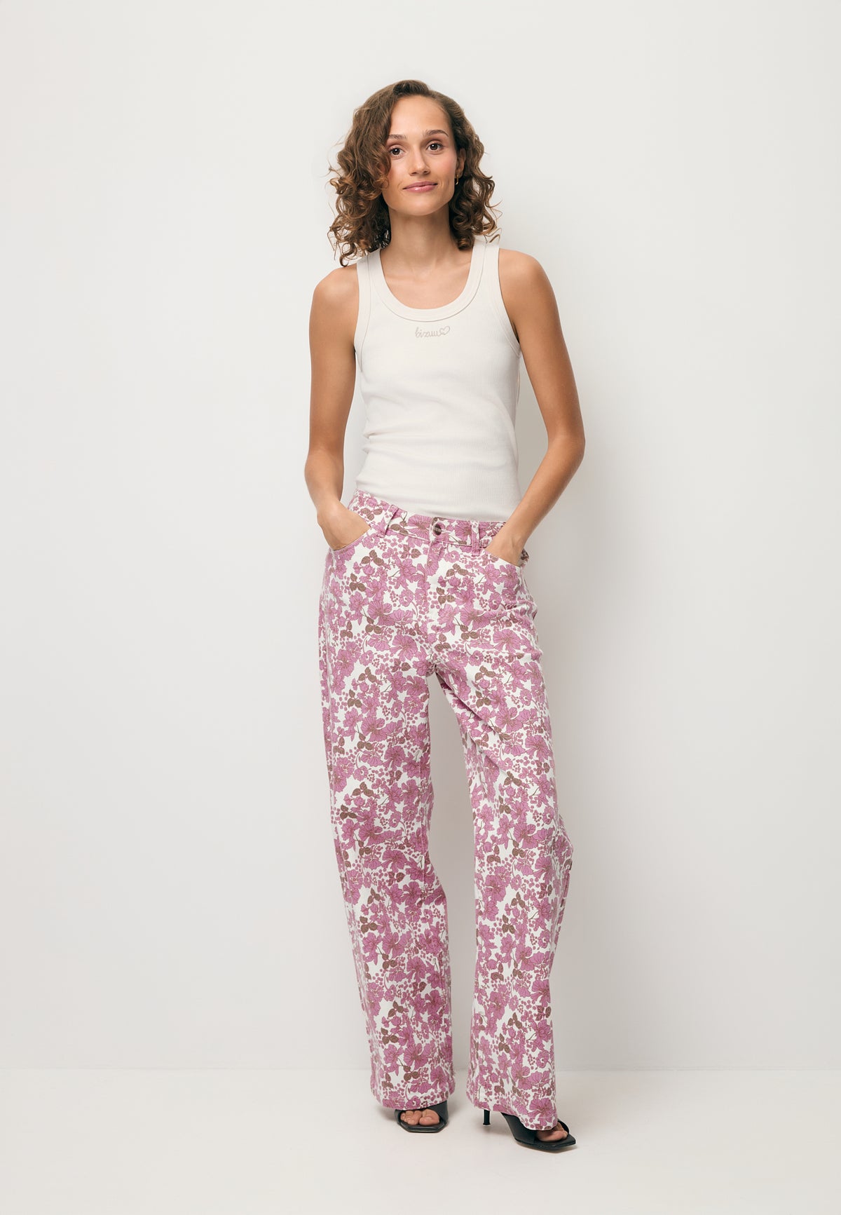 A model with her hands in the pockets of the DONY jeans in a floral print, wearing a white LOVIE top with a beige logo.