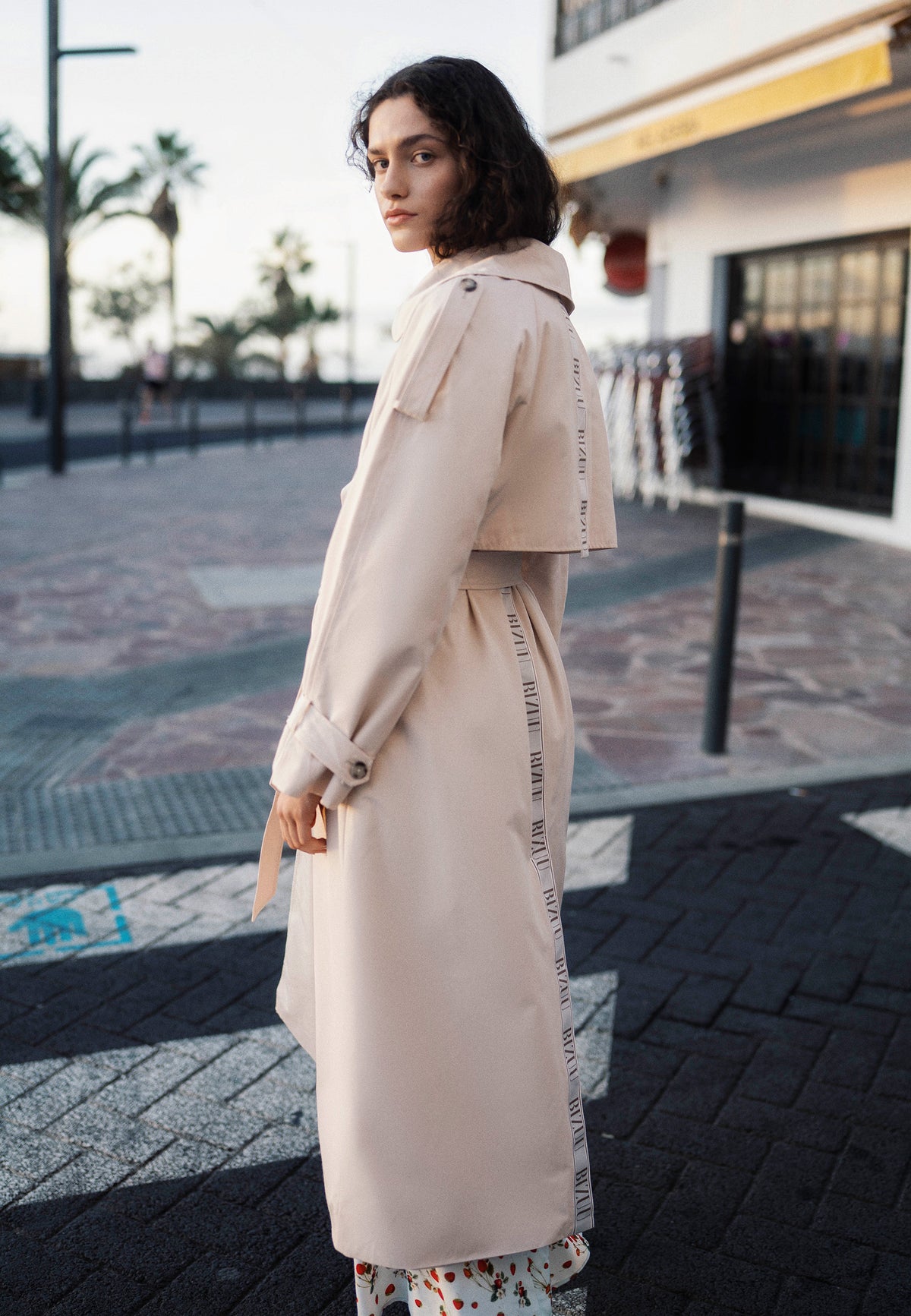 A model standing sideways at a pedestrian crossing, wearing a beige RACHELLA trench coat with a logo tape detail on the back.