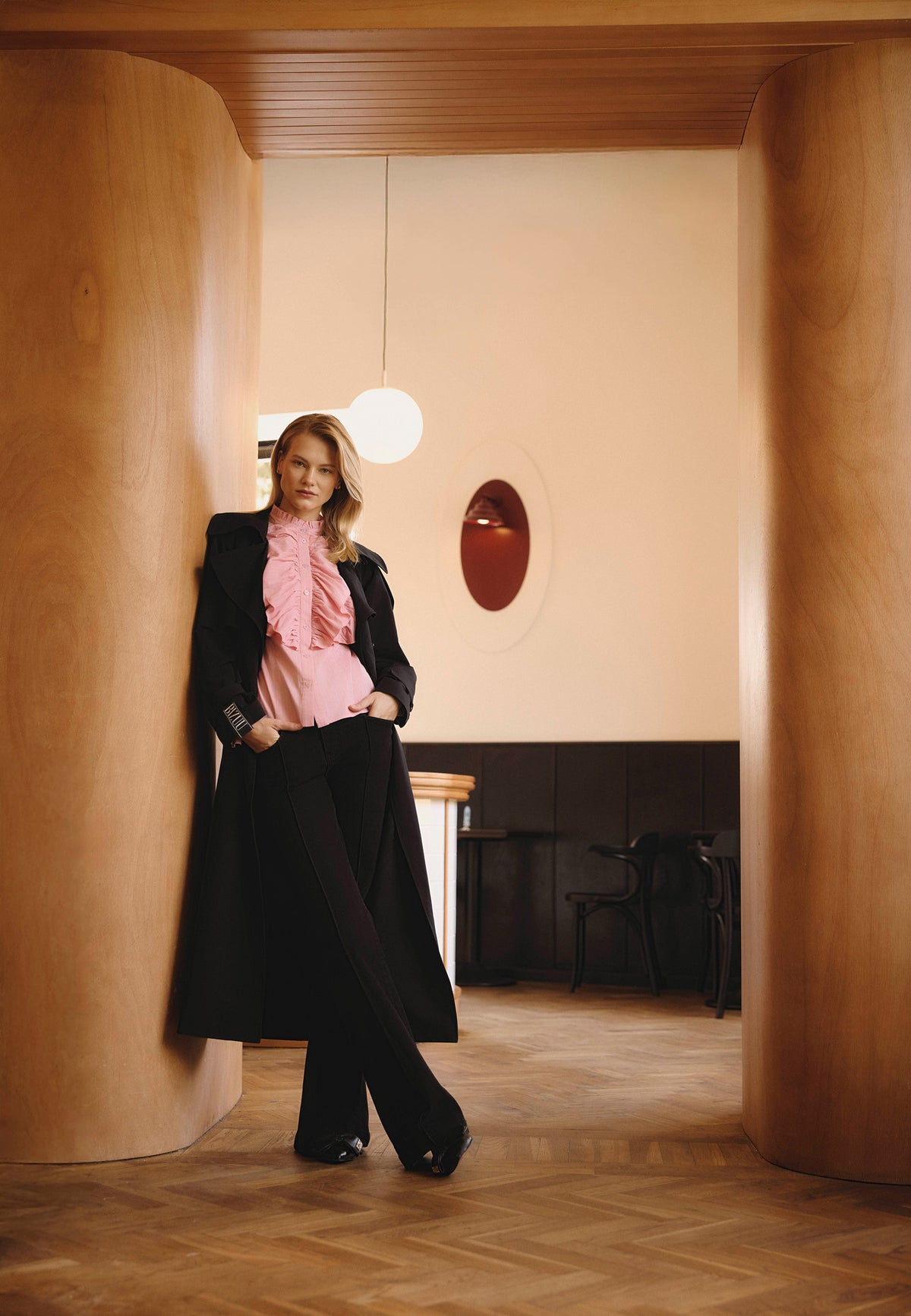A model in a café leaning sideways against a wooden wall, wearing a RACHELLA coat, SANNI shirt, and BELLINA jeans.