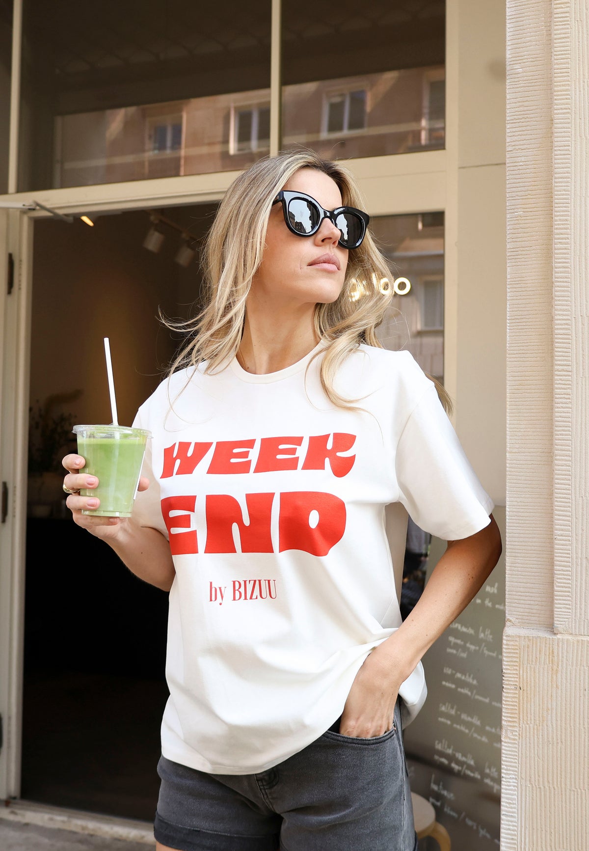 Close-up of the model with one hand in the pocket of MENORCA denim shorts holding a drink in her other hand. She is wearing a white T-shirt with red WEEKEND by Bizuu RALZA lettering.