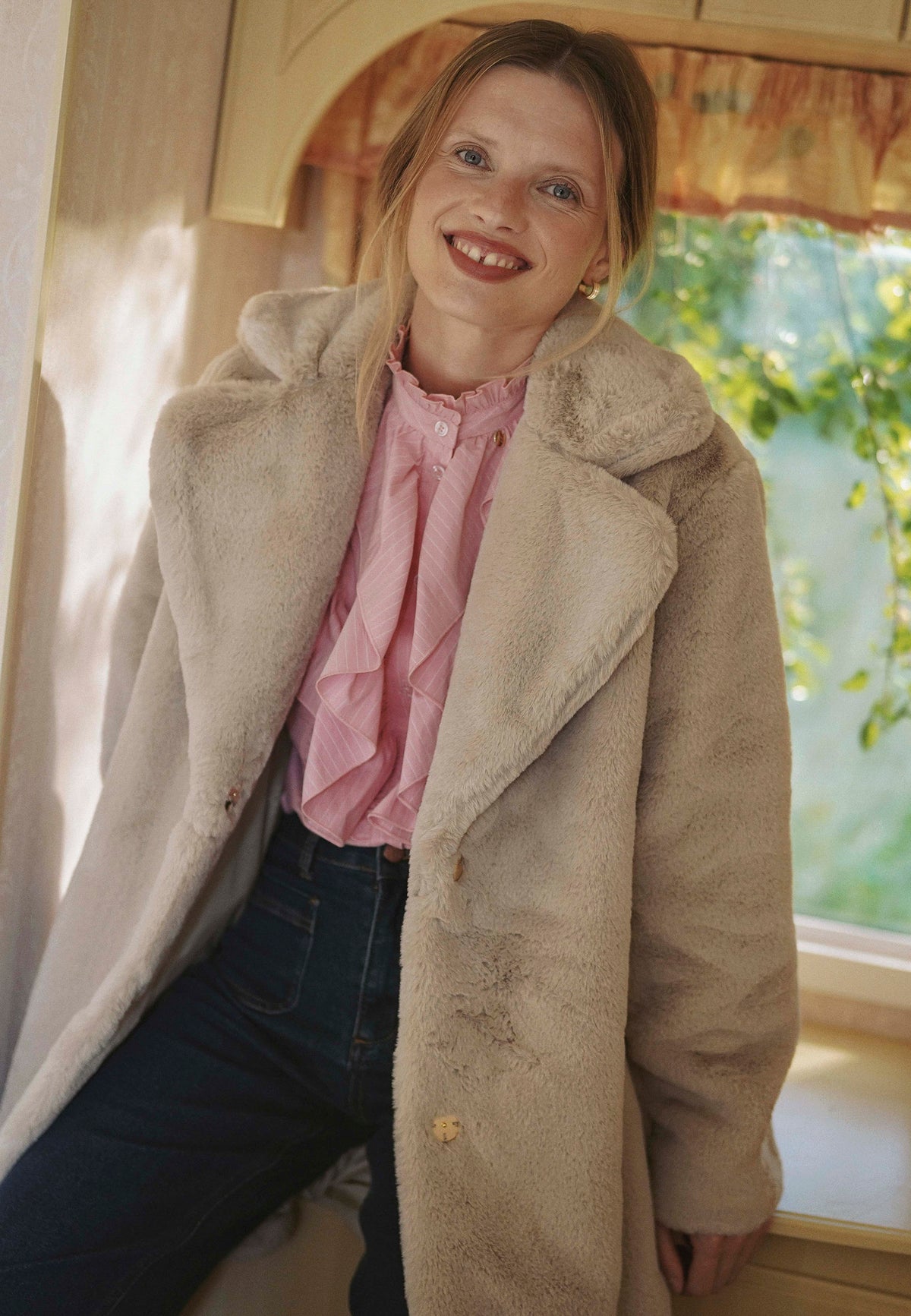 Model leaning against a wall, looking to the side, wearing the beige ROMA faux fur coat with embossed logo on the sleeve.