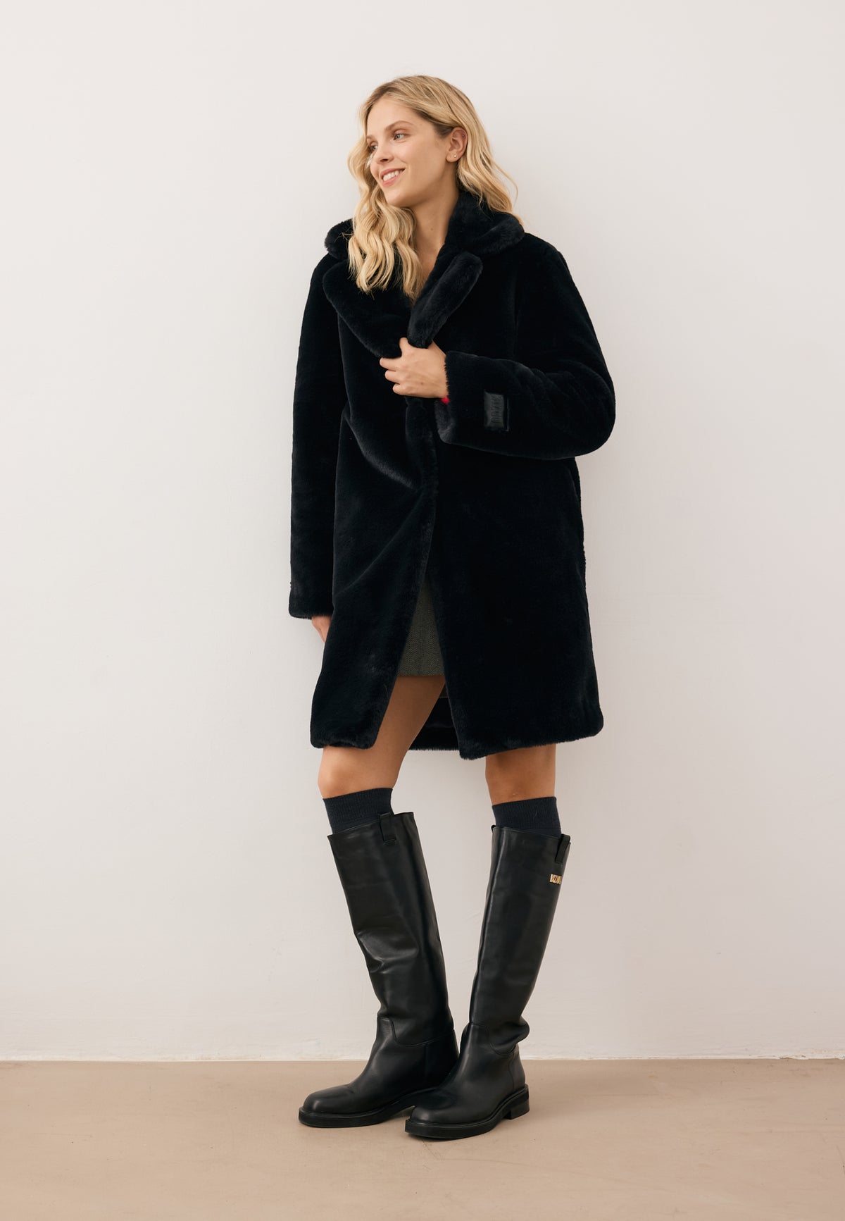 Model looking to the side, holding the collar of the black ROMA faux fur coat with embossed black sleeve logo, styled with CALYX black leather boots.