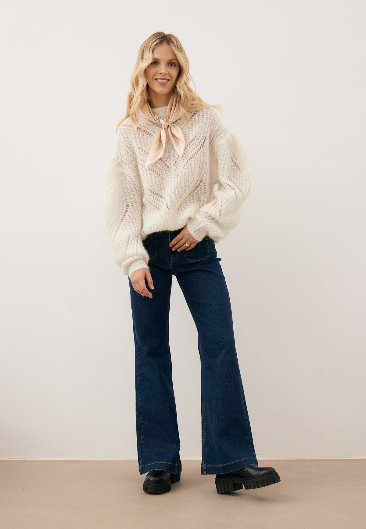 Smiling model wearing the cream ROSSO openwork sweater, DELPHI jeans, and the silk ARIA scarf.