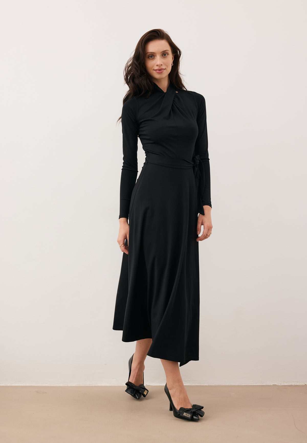 Smiling model taking a step in the black midi ROTTA dress with a decorative knot at the neckline.