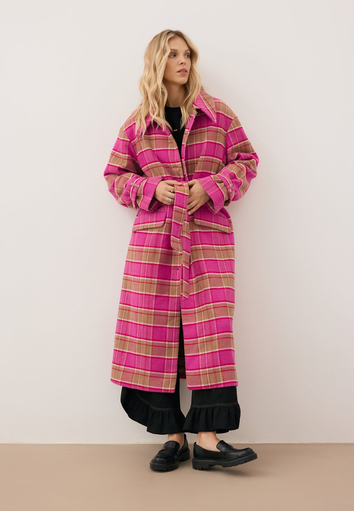 A model on a white wall wearing a pink WIVINA plaid coat, a SANNI shirt, and DELPHI pants.