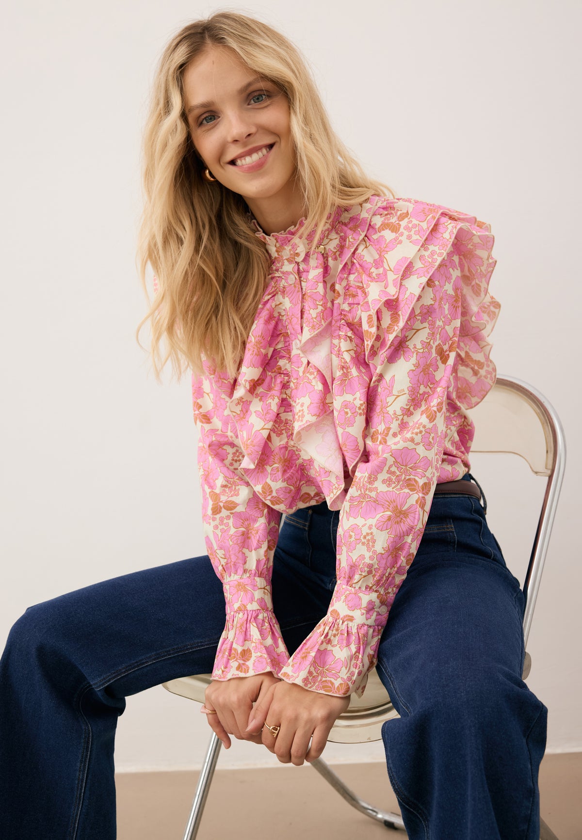 A model sitting on a chair wearing a floral SANNI shirt with a decorative collar and navy blue DELPHI jeans.