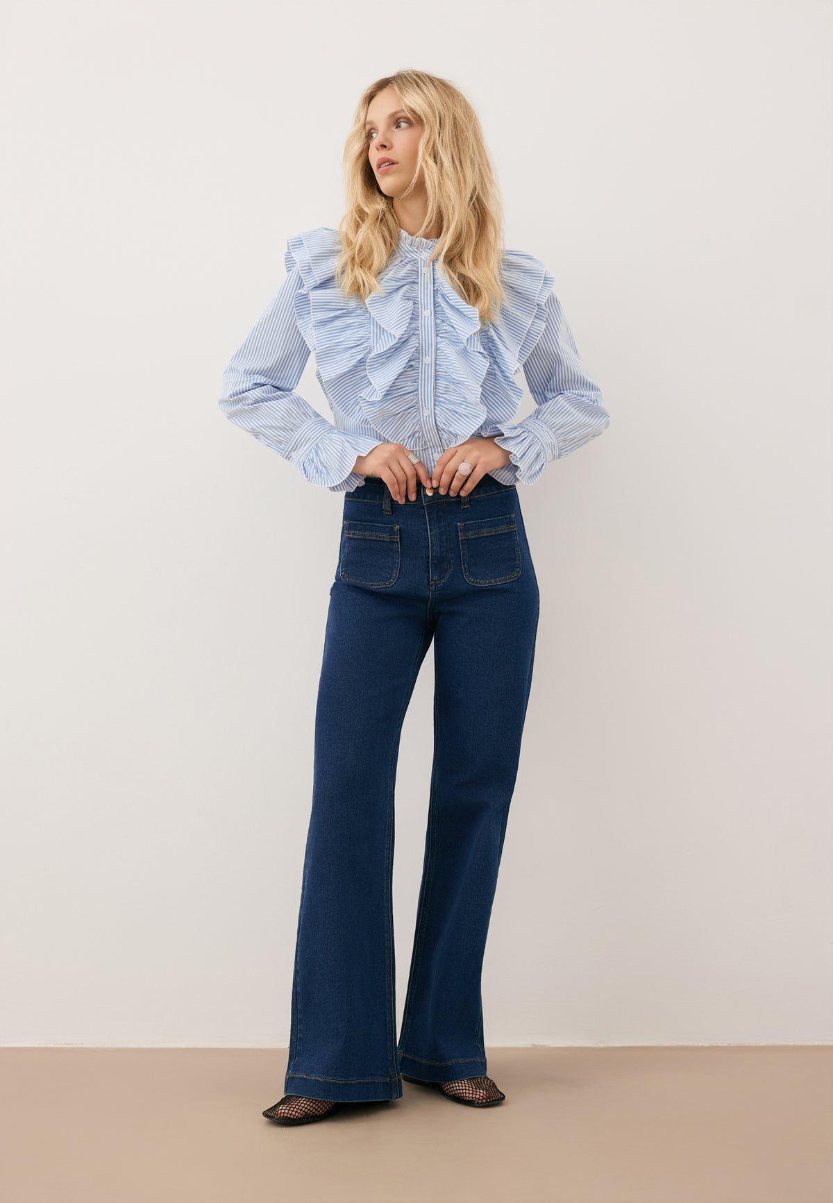 A model with her arms bent, wearing navy blue DELPHI jeans and a blue SANNI shirt with ruffles.