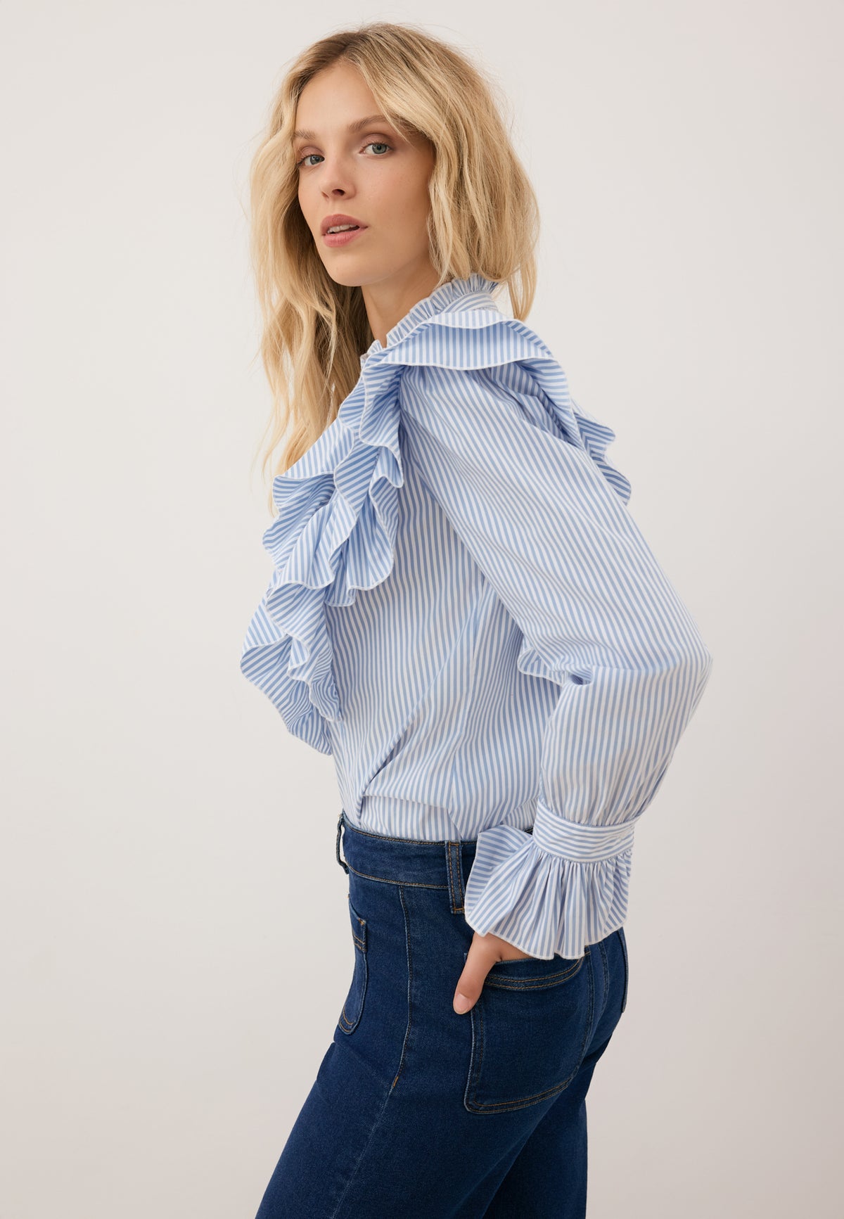 Close-up of a model standing sideways with her hands in the back pockets of DELPHI jeans. She is wearing a blue shirt with decorative frills SANNI.