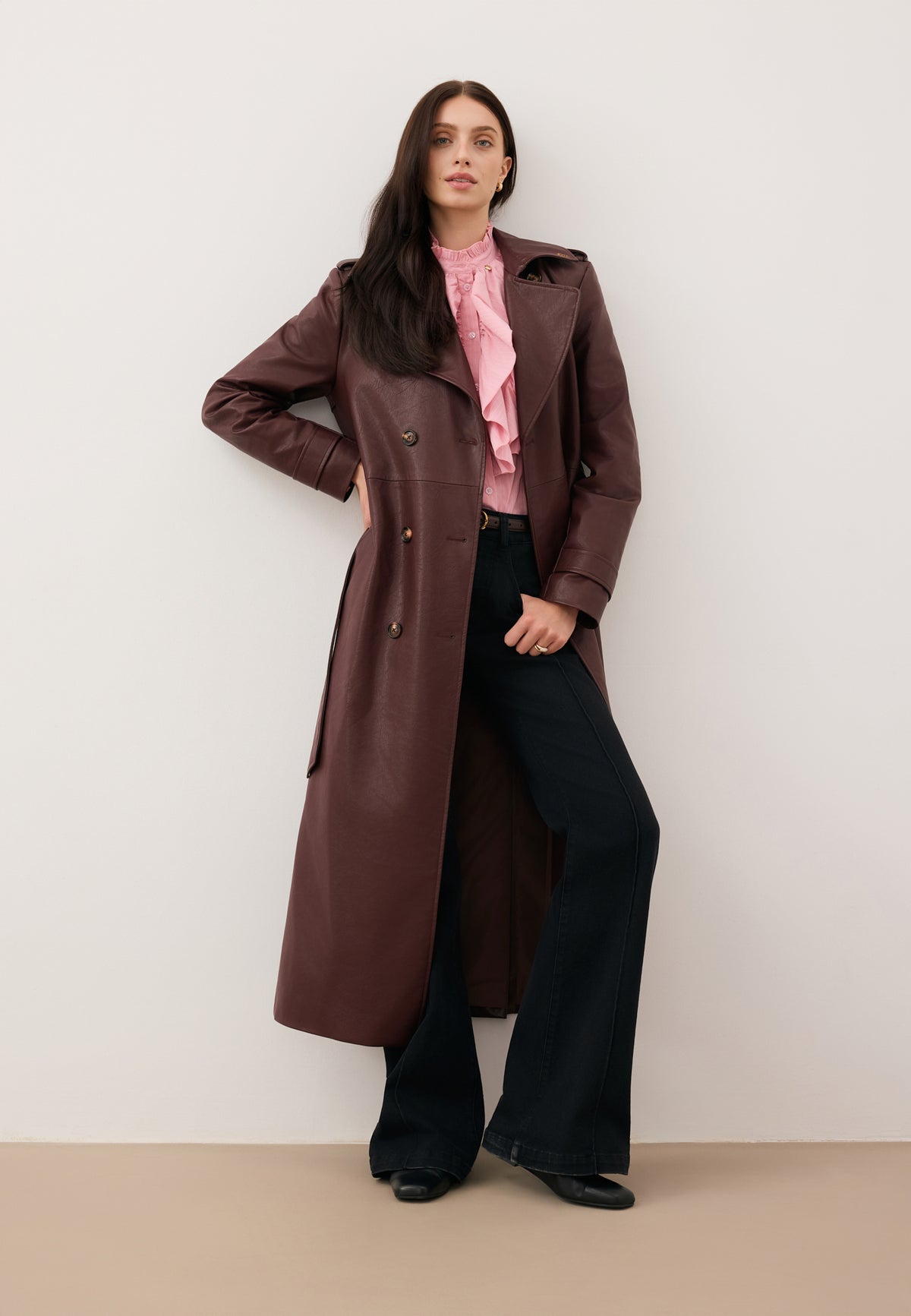 A model leaning against a wall with her hand on her hip, wearing a pink SANNI shirt, black BELINNA jeans, and a brown VENEKIA coat.