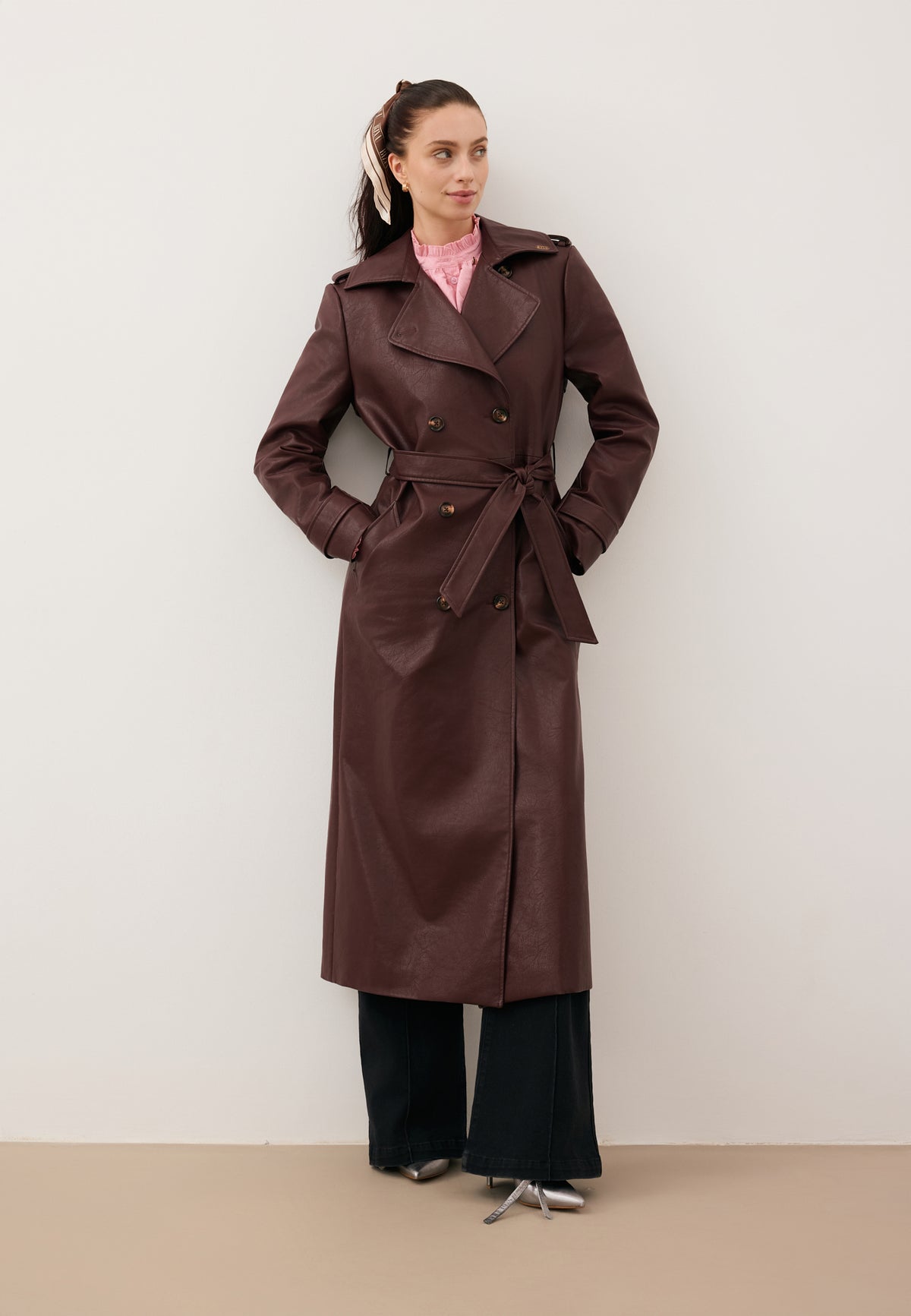 Model against a white wall wearing a brown VENEKIA coat tied at the waist and black BELINNA jeans.