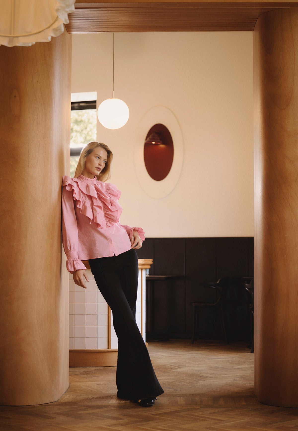 A model in a café leans against a wooden wall, standing with her legs crossed. She is wearing a pink SANNI shirt with ruffles and black BELLINA flare jeans.