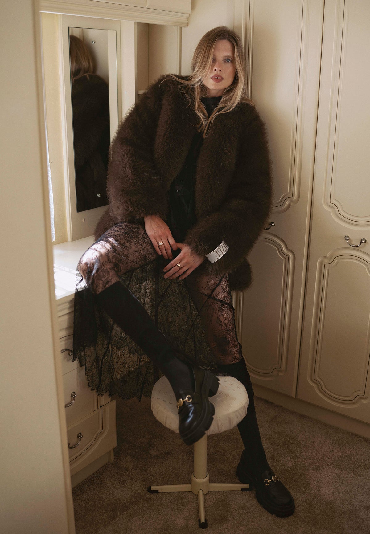 A model sitting on a desk wearing the SIENA brown long-haired faux fur coat and the FLACE lace skirt.