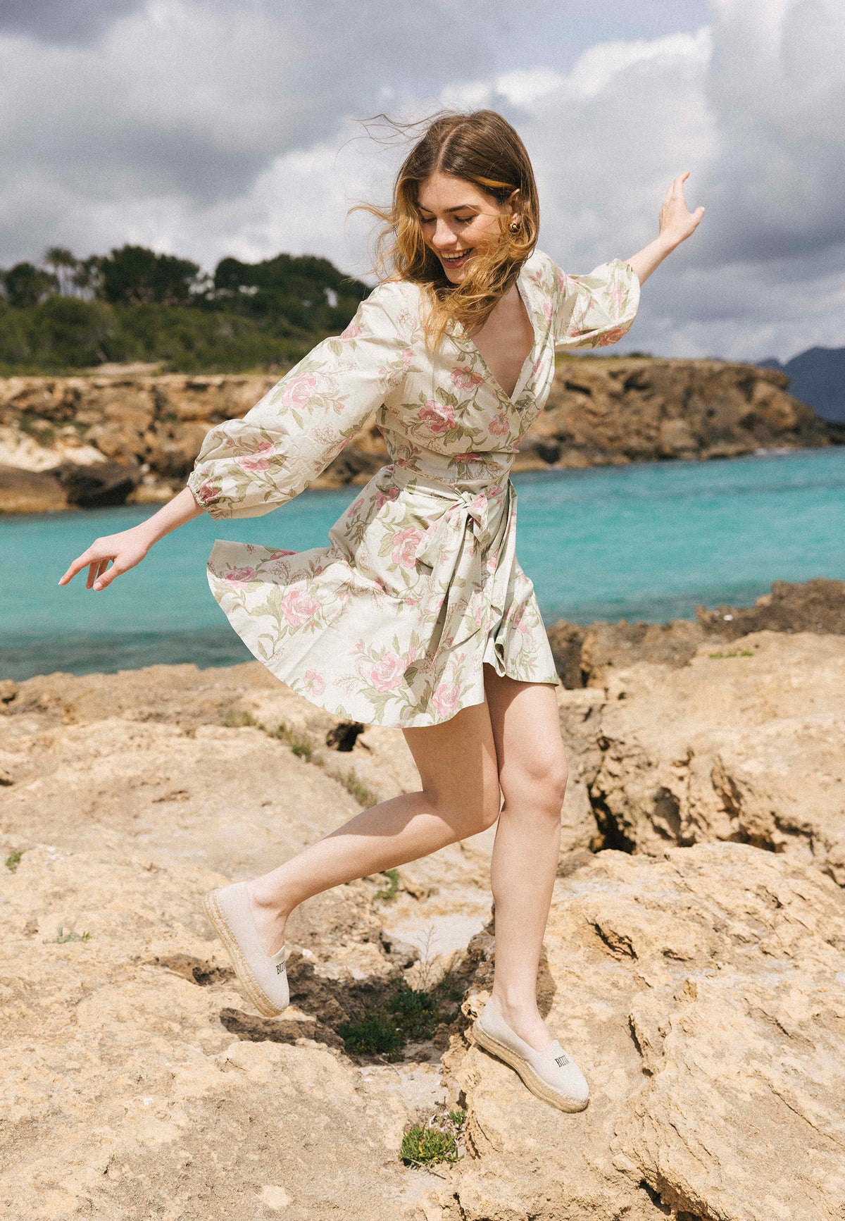A woman in a cream AMBROSIA mini dress with a pink floral print and 3/4 sleeves, leaning against rocks, shields her eyes from the sun.