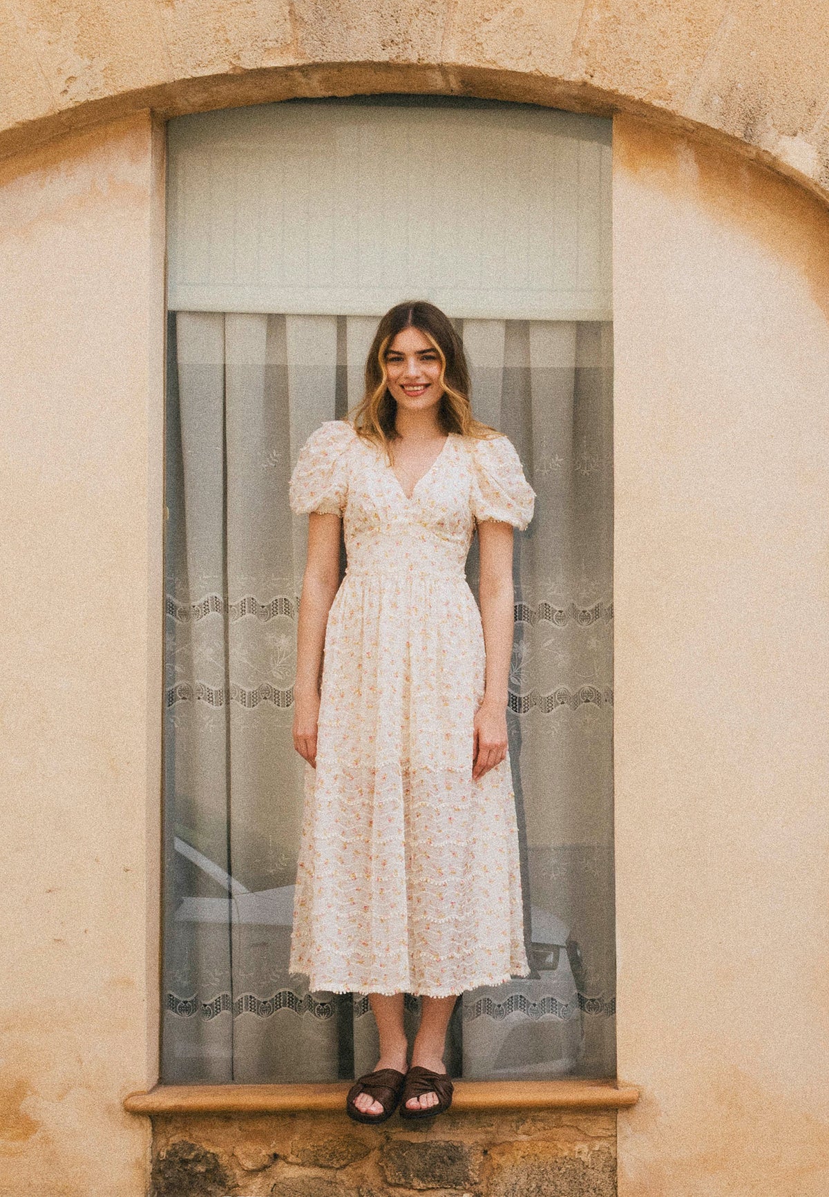 A smiling woman in a romantic midi dress in pastel flowers with FIFFI buffets, stands on the window sill with a curtain.