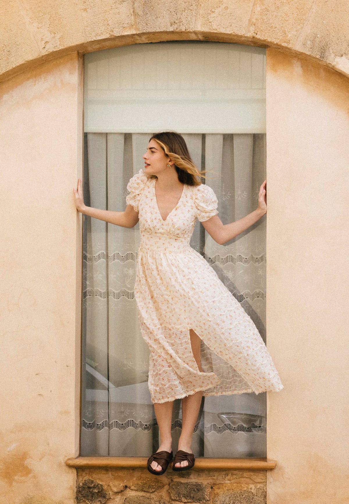 A woman in a light midi dress with tiny FIFFI flowers stands in a window recess, holding her sides.