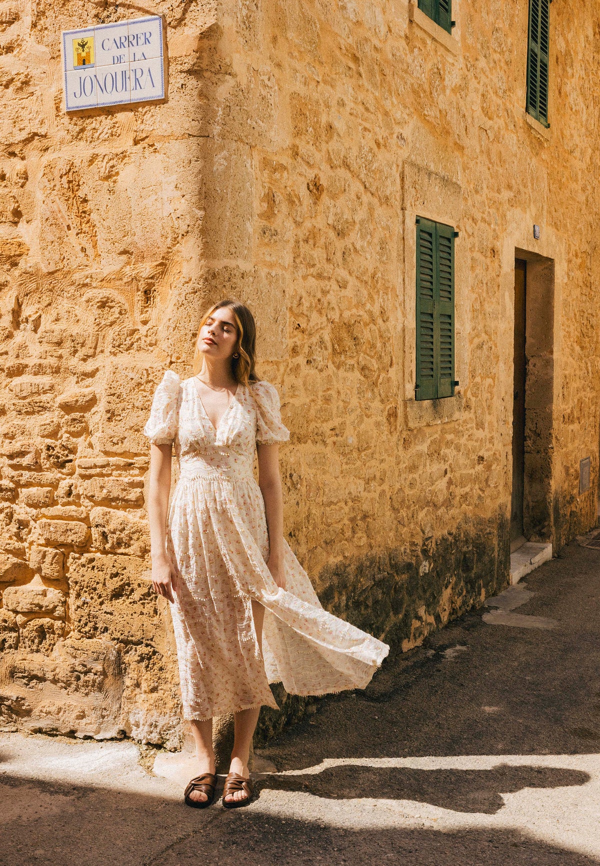 A model in a dress in small flowers with a slit on the leg of FIFFI, standing in the sun by the stone wall of a building.